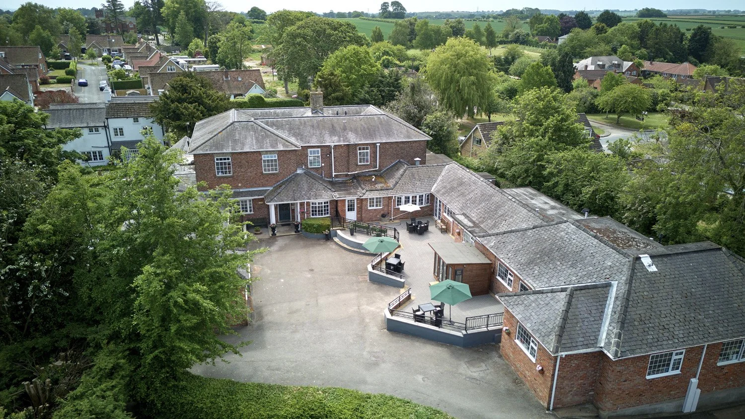 Aerial view of a large brick house with multiple roofs, surrounded by greenery and trees, with a spacious backyard roofed outdoor patio area with furniture, umbrellas, and shrubs, in a suburban neighborhood.