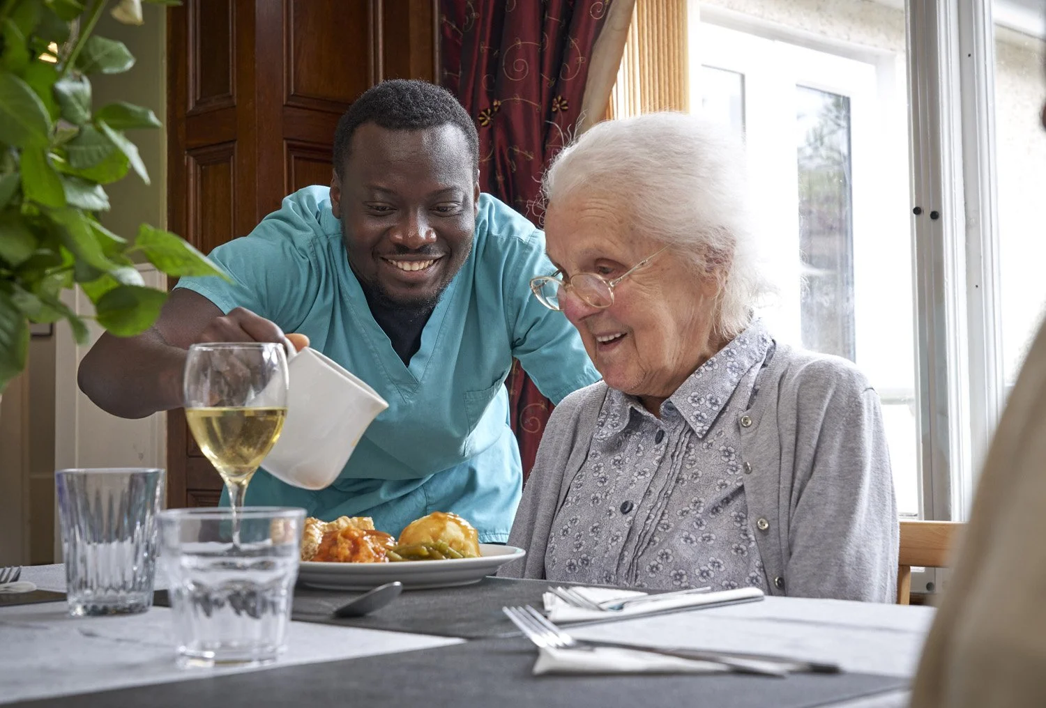 A man in medical scrubs serves food to an elderly woman at a dining table, both smiling.