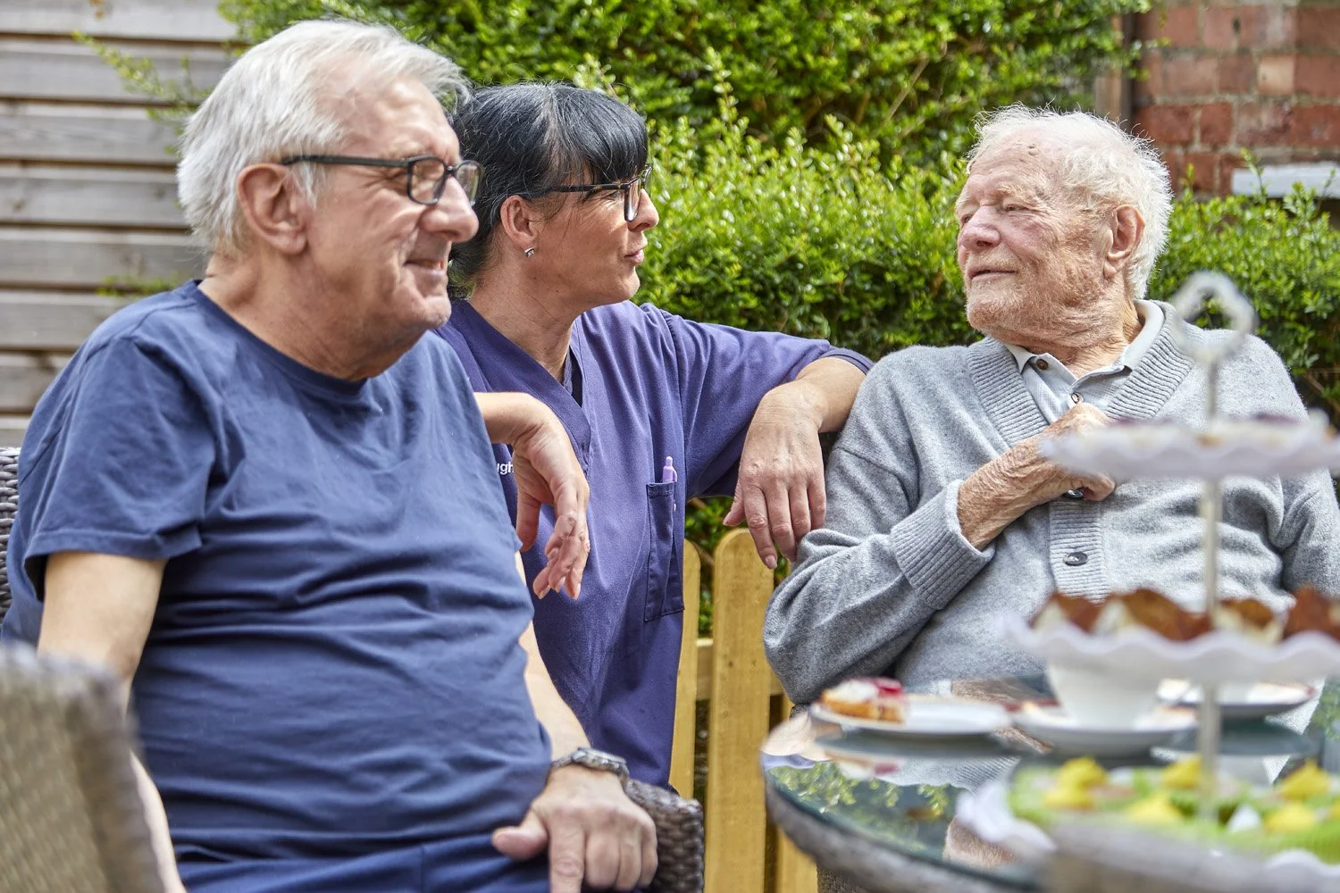 Three elderly people sitting at an outdoor table with a tiered tray of desserts, chatting and enjoying each other's company.