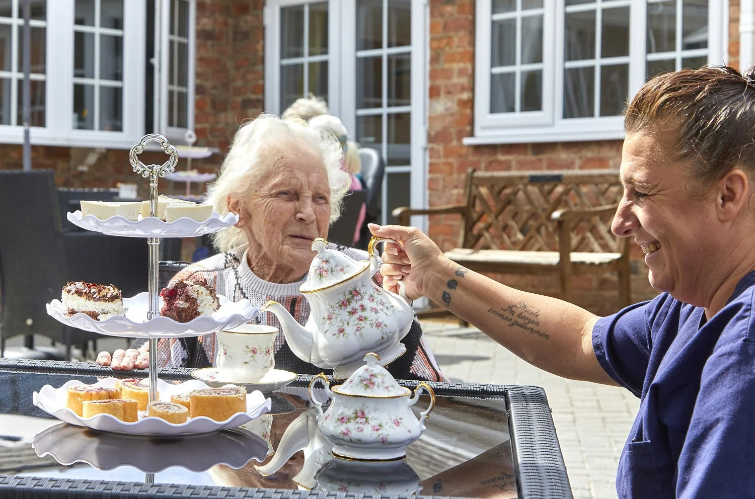 A woman hands a teapot to an elderly woman during a tea party outside, with a three-tiered tray of desserts on the table.
