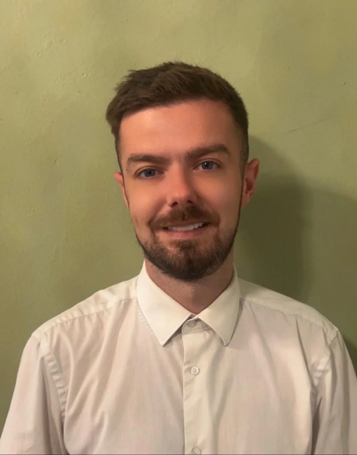A young man with short brown hair, a beard, and mustache, wearing a white dress shirt, standing against a light green wall.