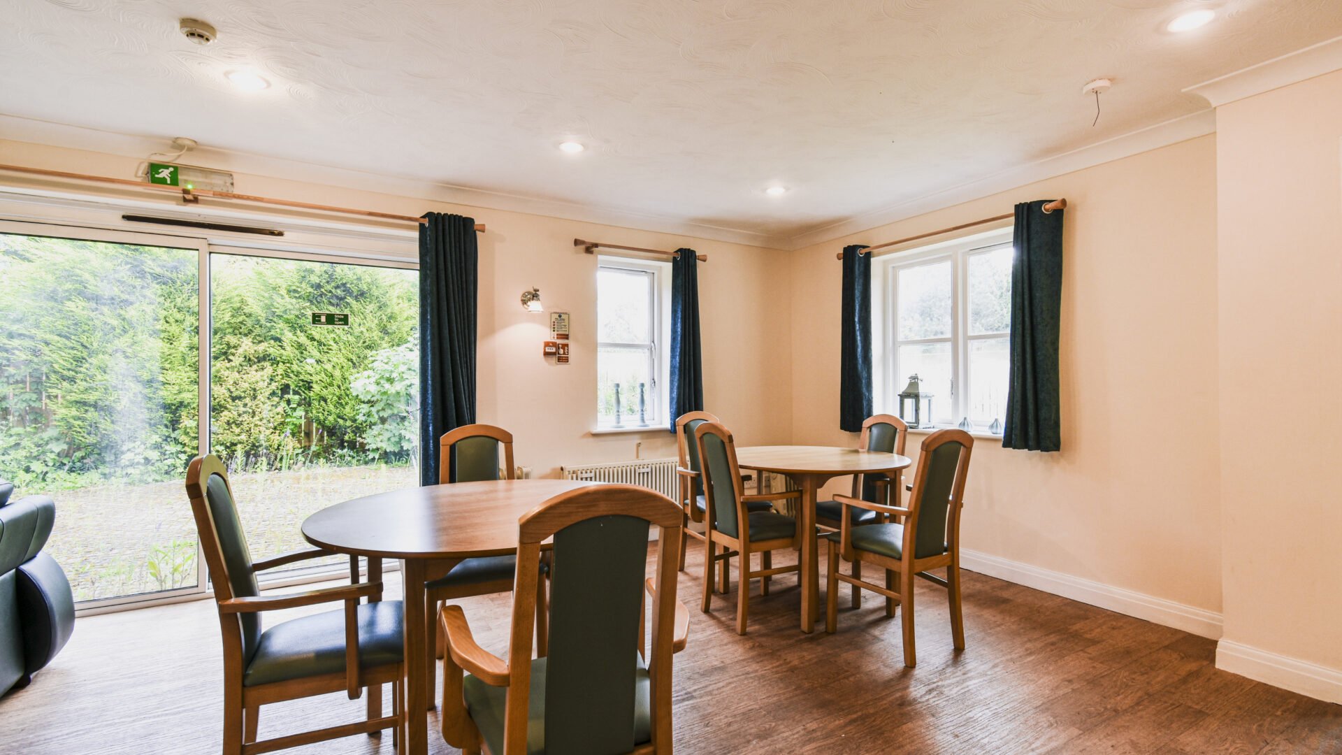 Empty dining area with wooden tables and chairs, large window with blue curtains, and sliding glass door overlooking a green outdoor space.