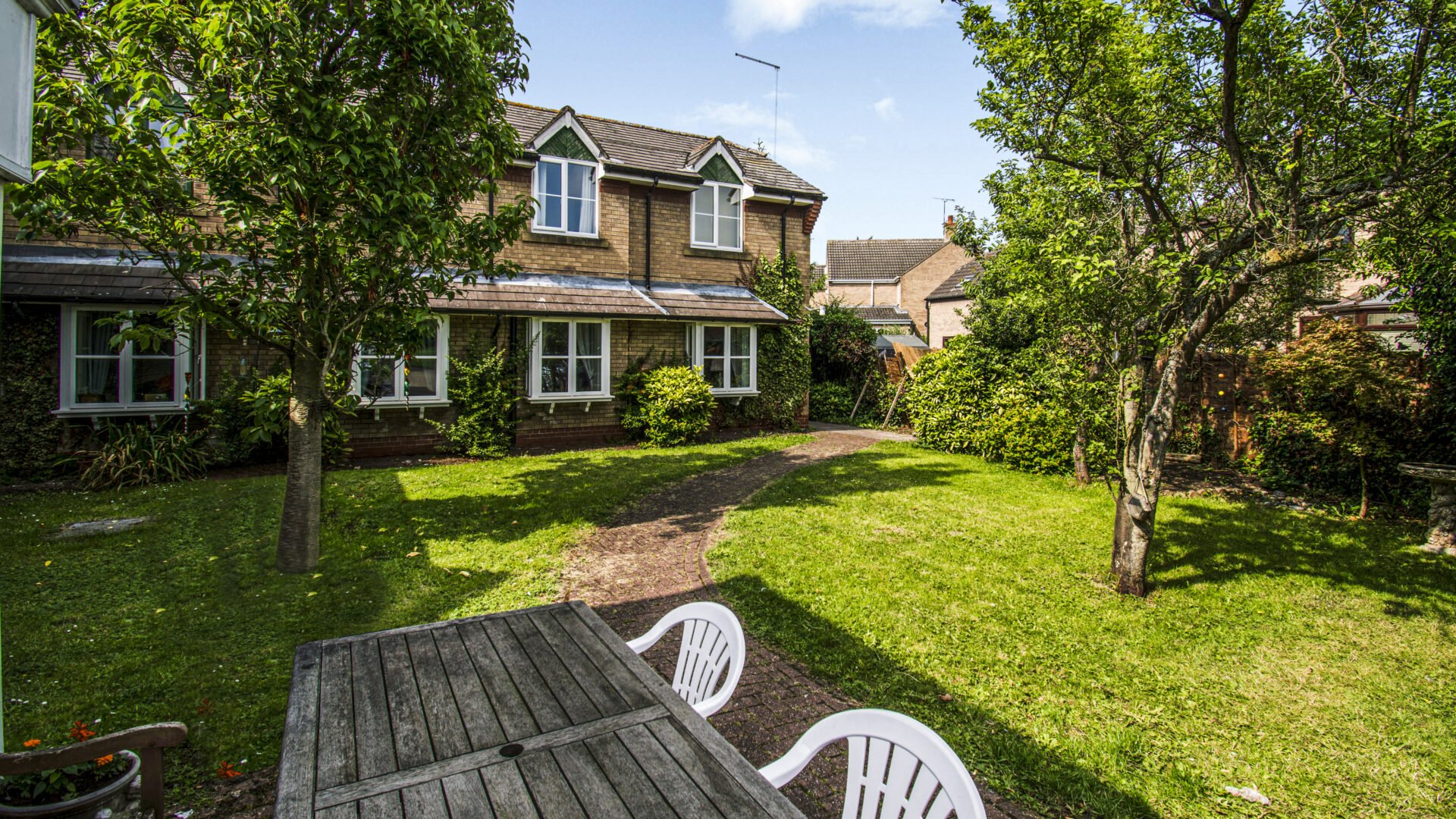 Backyard garden with green grass, trees, and a brick pathway, view of neighboring houses and a wooden table with white chairs in the foreground.
