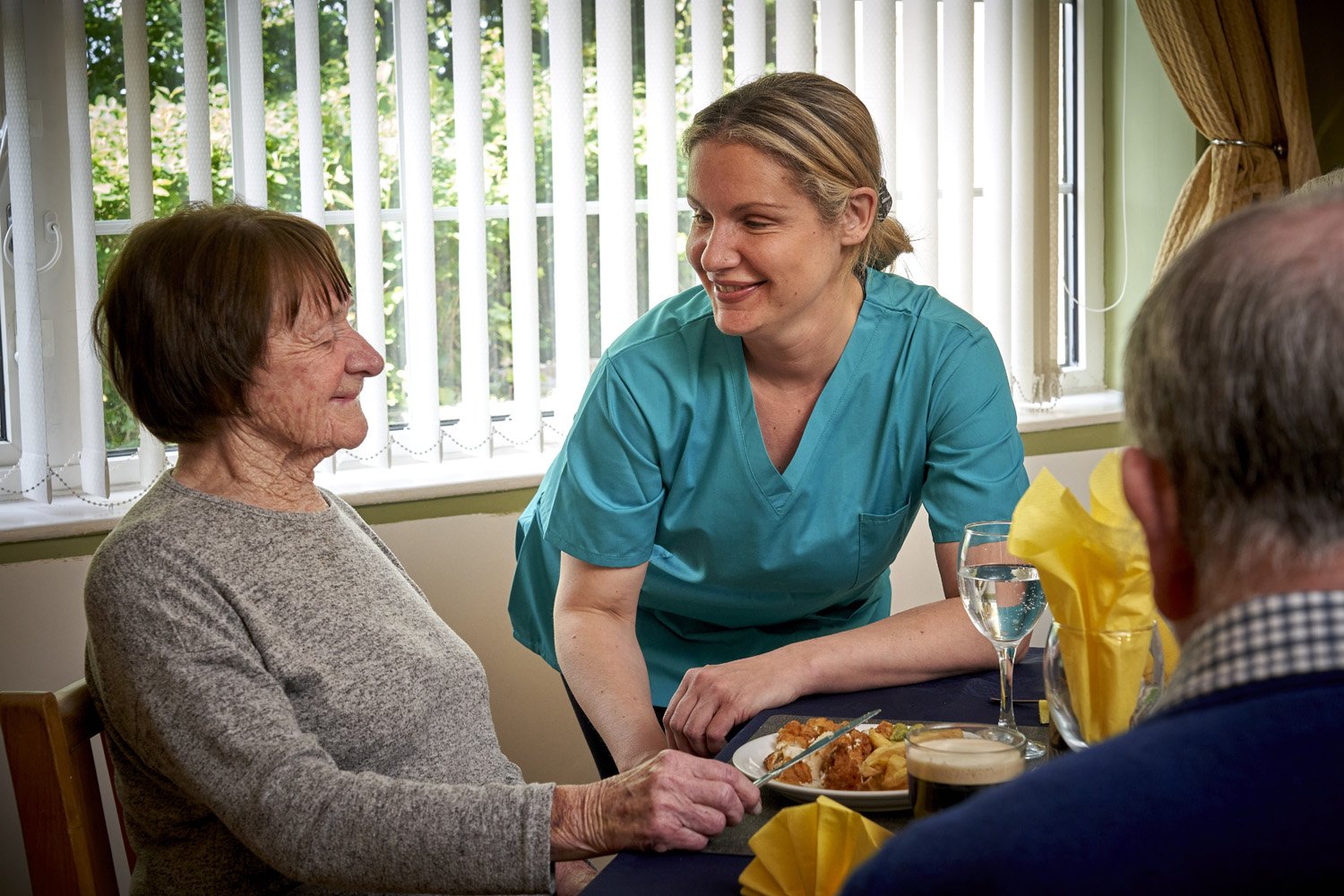A nurse smiling and talking to an elderly woman at a table, with other elderly individuals present, in a dining room with large windows and natural daylight.