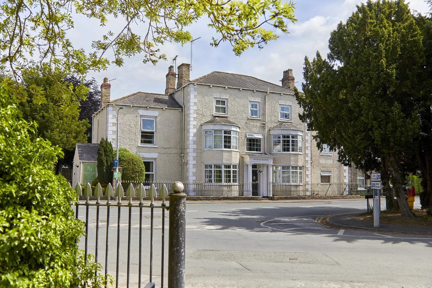 A multi-story, pale-colored residential building with bay windows, surrounded by trees and shrubs with a small yard and a fence in front, located on a corner street with road signs nearby.