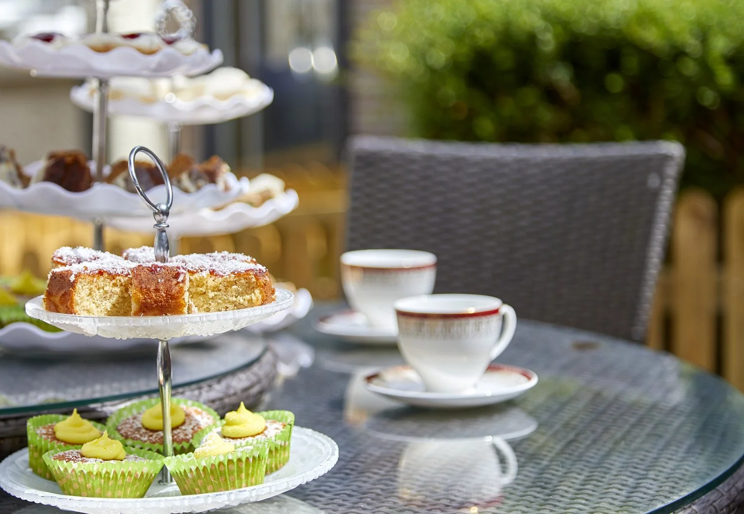 Outdoor table with a tiered tray of assorted finger foods and desserts, two cups of tea or coffee, and a wicker chair in the background, surrounded by greenery.