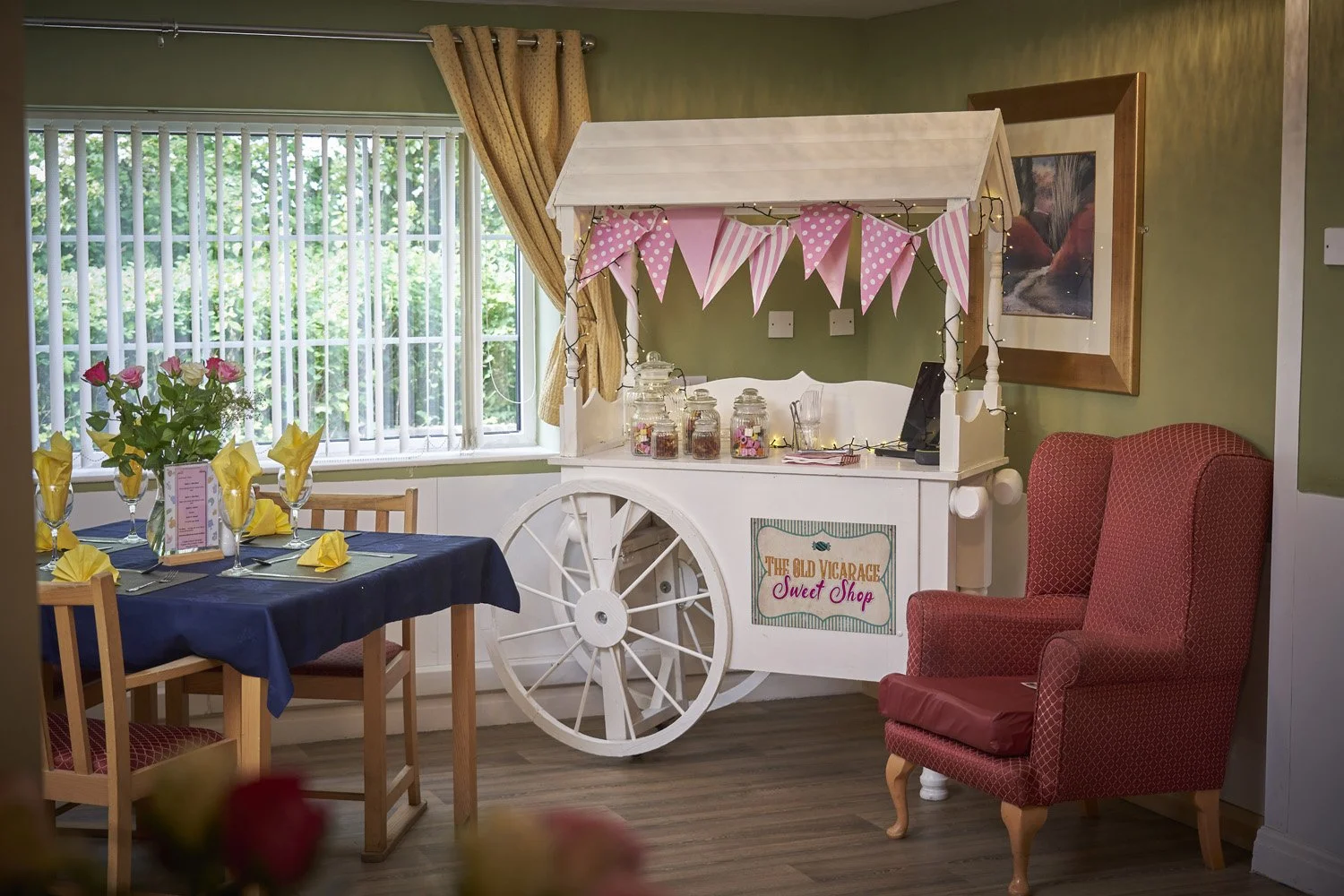 Decorative indoor scene featuring a vintage white candy cart with pink pennant banners, jars of candies, and a sign that reads 'The Old Vicarage Sweet Shop.' To the left, a dining table with a navy tablecloth, yellow napkins, glasses, and a vase of p