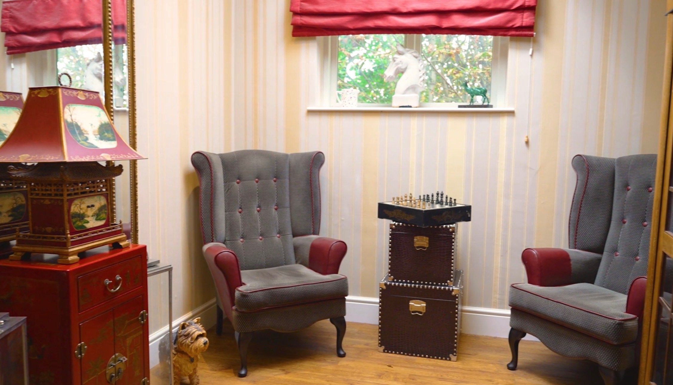 A cozy room with two high-backed armchairs, a small chess table between them, a red cabinet with decorative items, a table lamp, a small dog sitting on the floor, and a window with a white horse and a green sculpture outside.