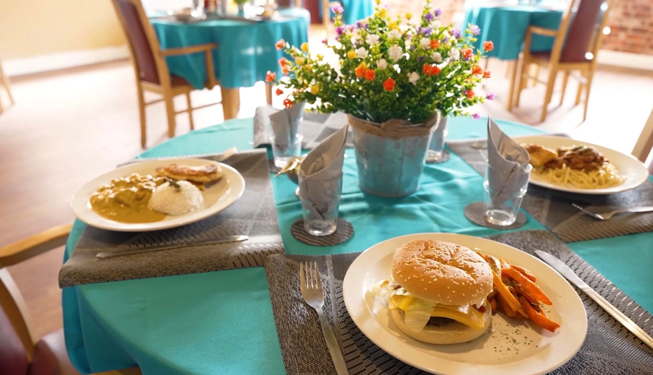 A dining table set with plates of food, including a burger with sweet potato fries, a plate of rice with curry and chicken, and a plate of spaghetti with meat sauce. There is a large floral centerpiece with colorful flowers in the middle, and the tab
