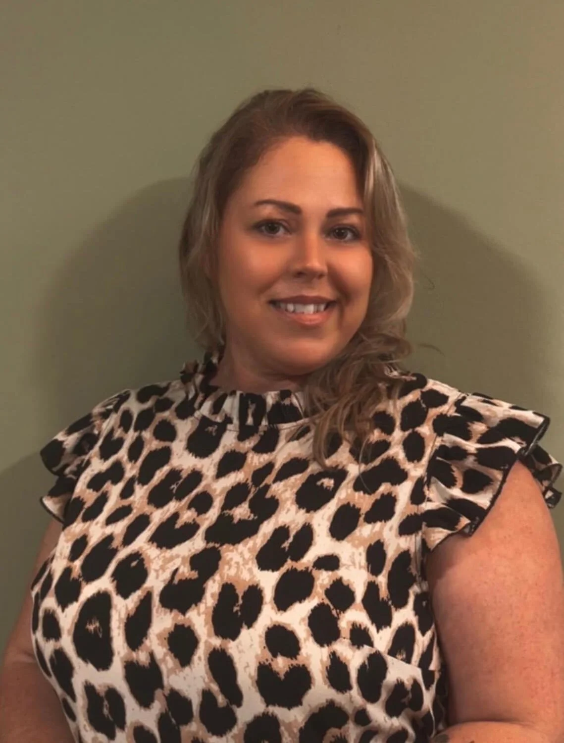 A woman with shoulder-length light brown hair smiling, wearing a sleeveless leopard print top, standing against a plain green wall.