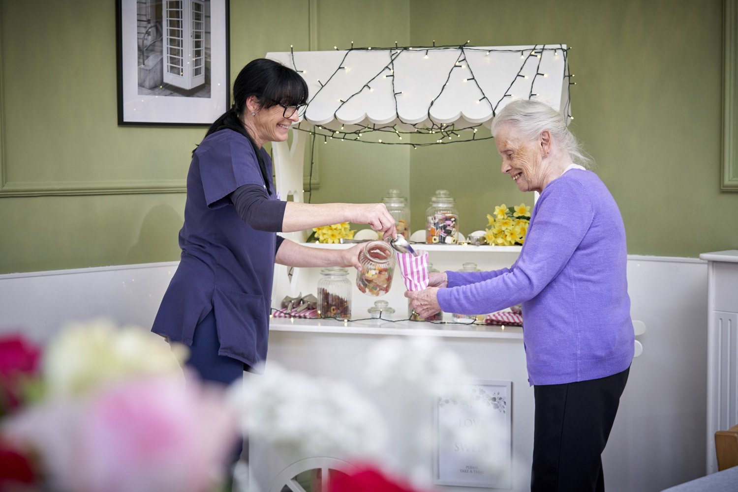 A young woman in purple scrubs helps an elderly woman in a purple sweater at a candy table, smiling and sharing a moment.