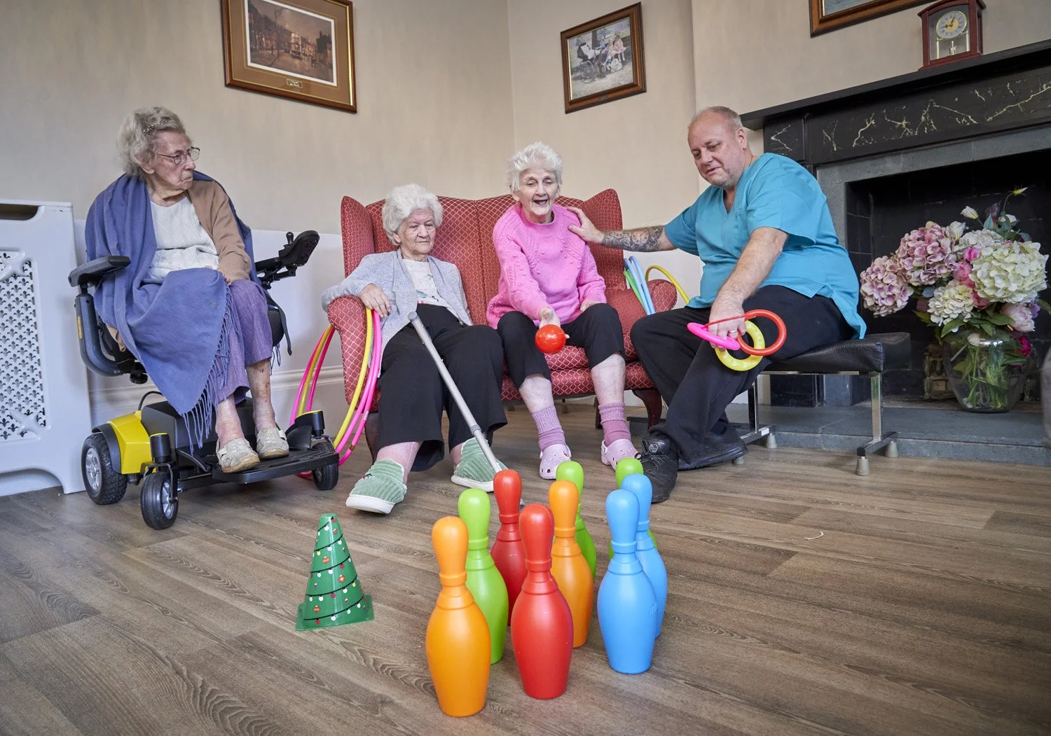 Four elderly women and a male caregiver playing a game with colorful bowling pins and balls in a cozy living room with paintings and flowers.