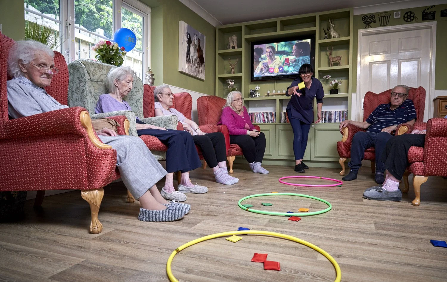 Elderly people sitting in a living room observing a woman playing a ring toss game with large colorful hoops and small red, blue, and yellow discs, while watching a television mounted on a green wall.