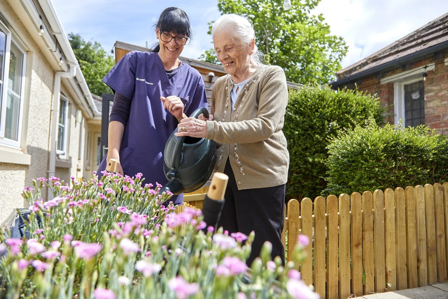 An elderly woman watering flowers in her garden while a caregiver supervises outdoors on a sunny day.