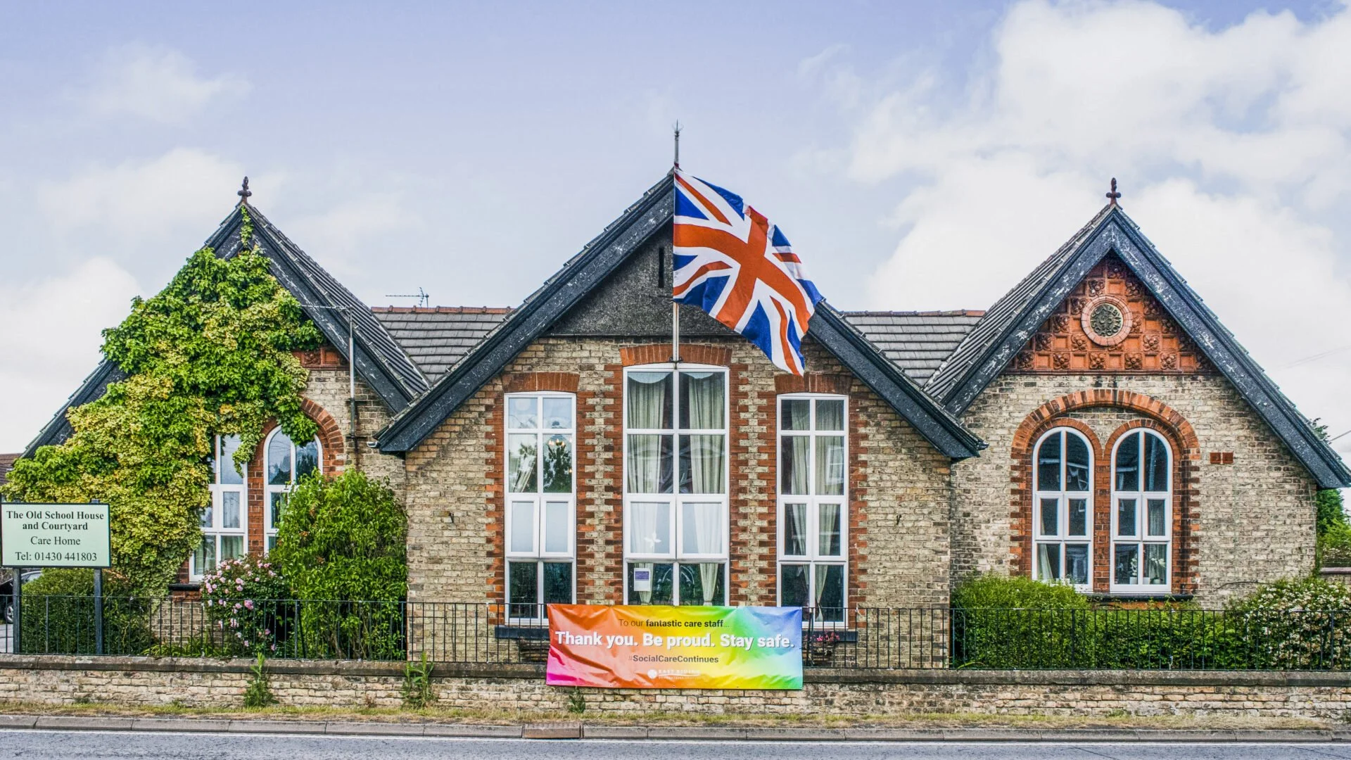 A brick building with large windows and a central flagpole displaying the Union Jack flag; there is a rainbow-colored banner at the front reading 'Thank you. Be proud. Stay safe.' and a green sign on the left indicating it is 'The Old School House an