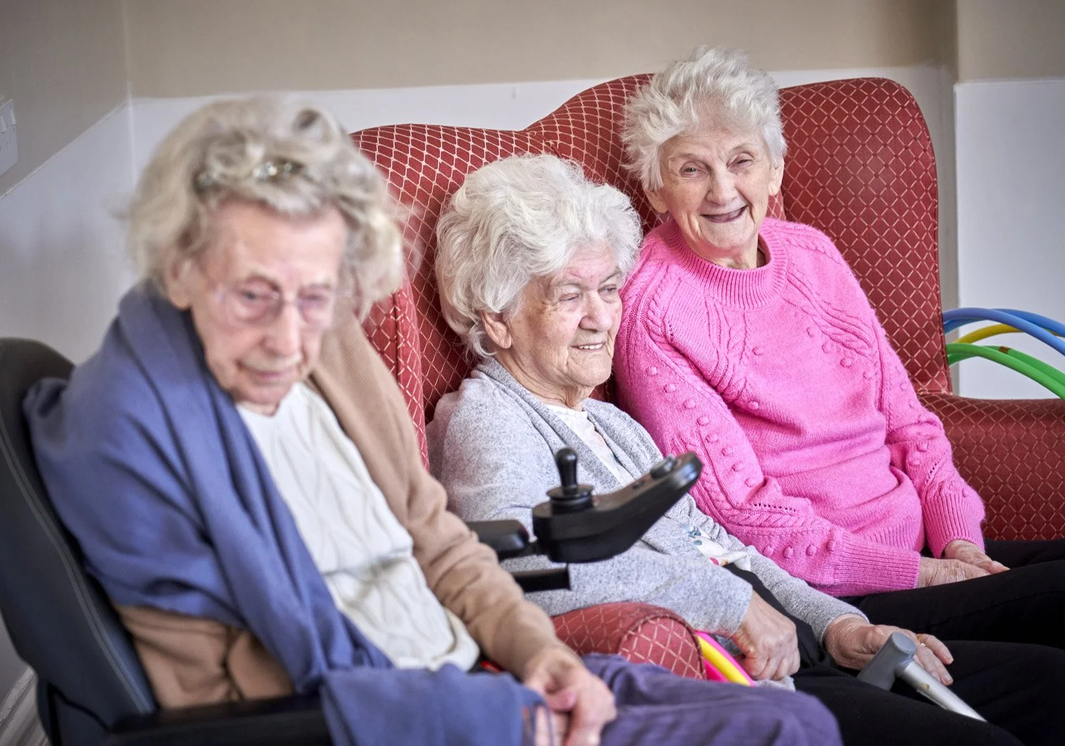 Four elderly women sitting together in a care facility, some using mobility aids, smiling and enjoying each other's company.