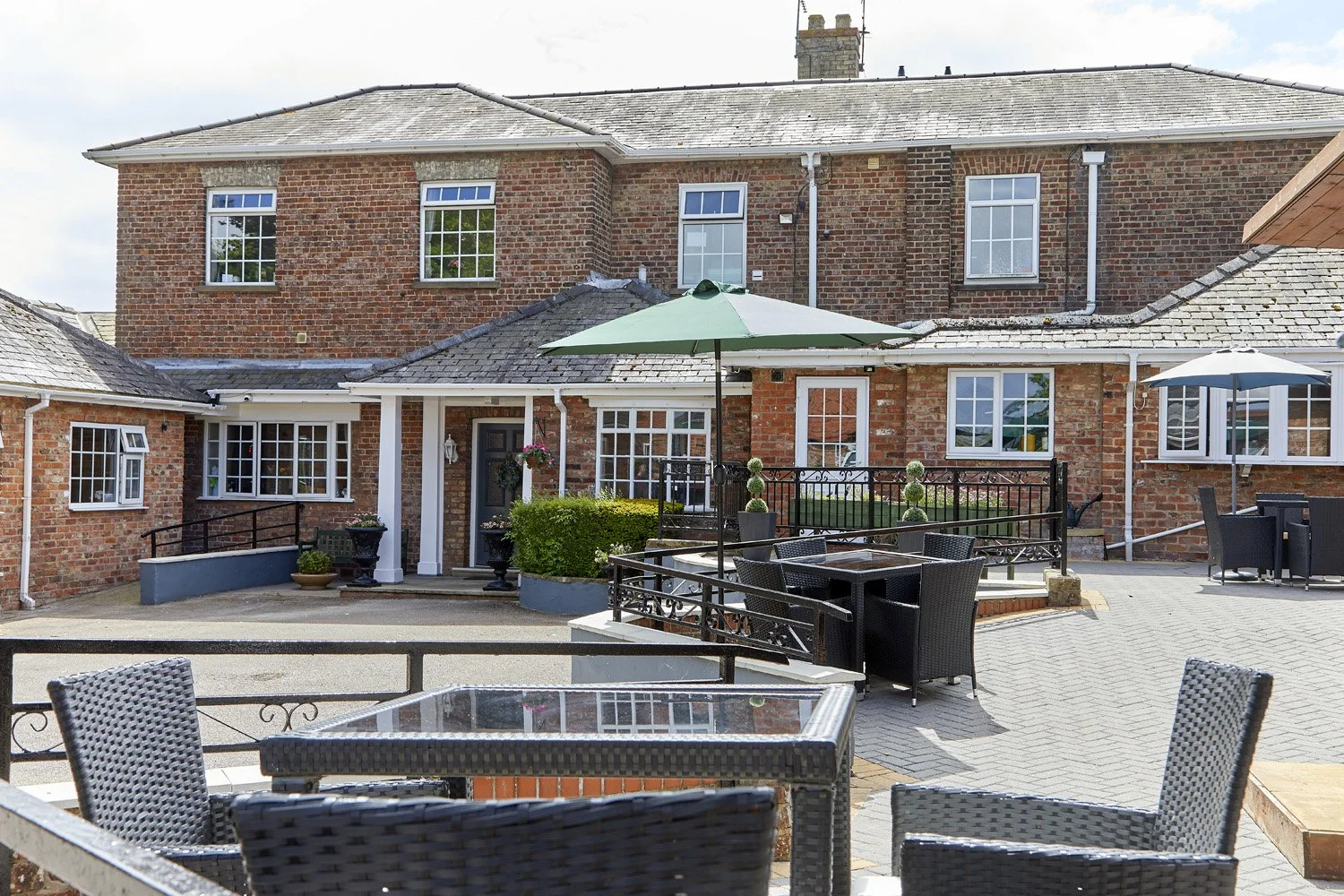 A spacious brick backyard patio with outdoor furniture, umbrellas, potted plants, and a view of a large brick house with multiple windows.