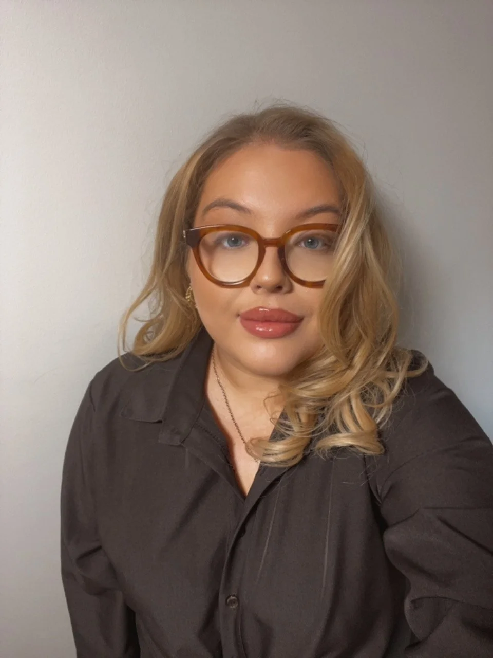 A woman with curly blonde hair and glasses taking a selfie against a plain wall.