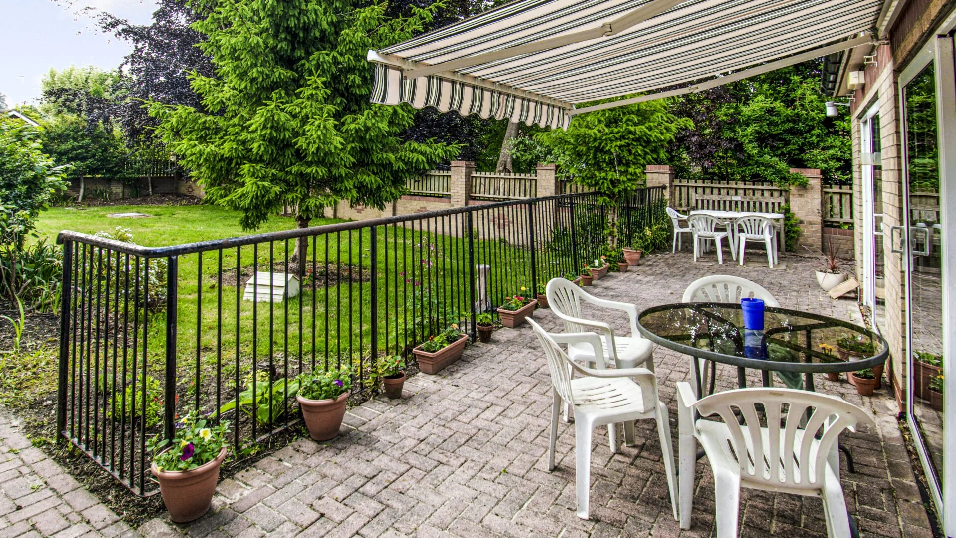A backyard patio with white plastic chairs around a glass table, potted plants, a metal fence, and a grassy lawn with trees and a brick wall in the background.
