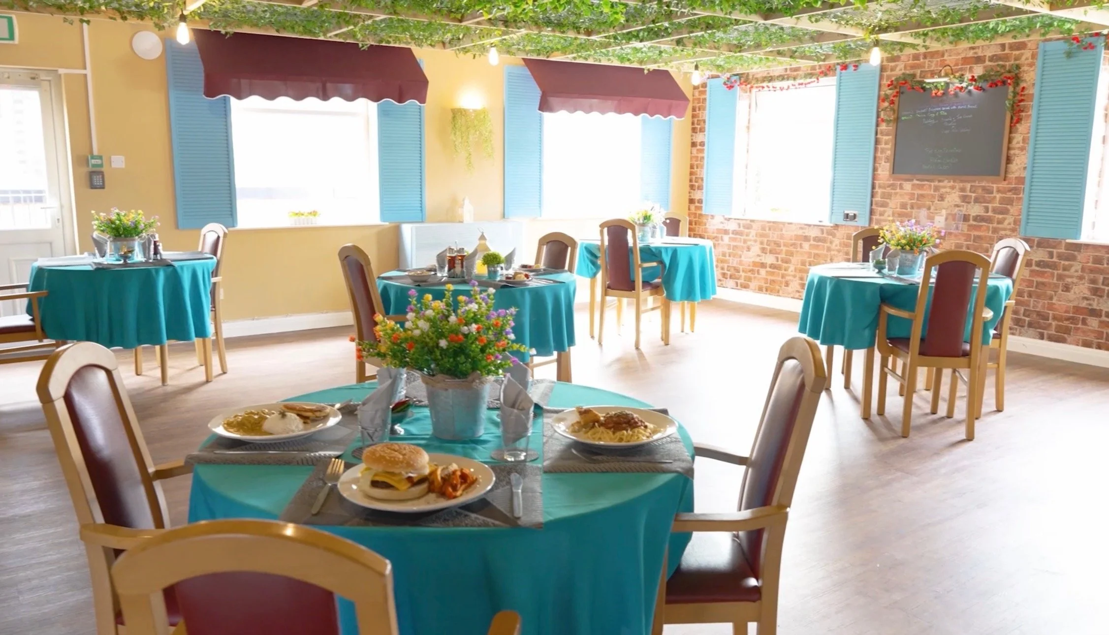A bright, cozy restaurant interior with turquoise tablecloths, floral centerpieces, and blue shutters on windows. There are several tables with plates of food, including burgers and pasta, set for dining. The room features a brick wall with a blackbo