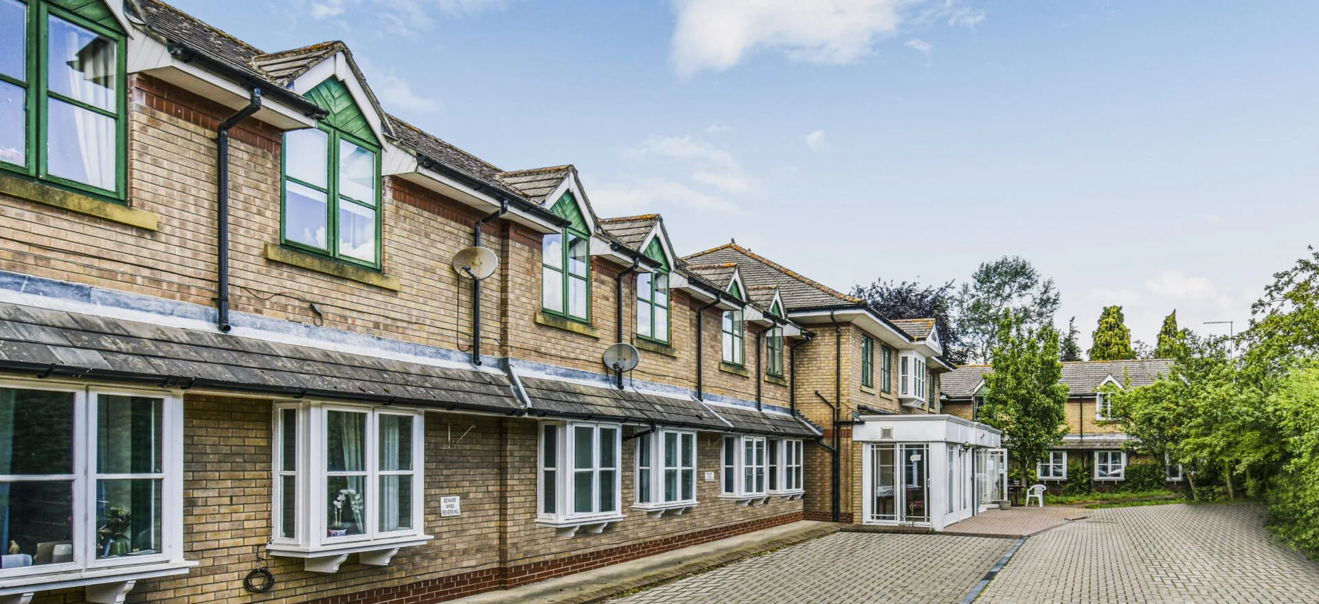 Residential apartment building with brick exterior, multiple windows with green trim, satellite dishes, and a paved courtyard surrounded by trees under a partly cloudy sky.