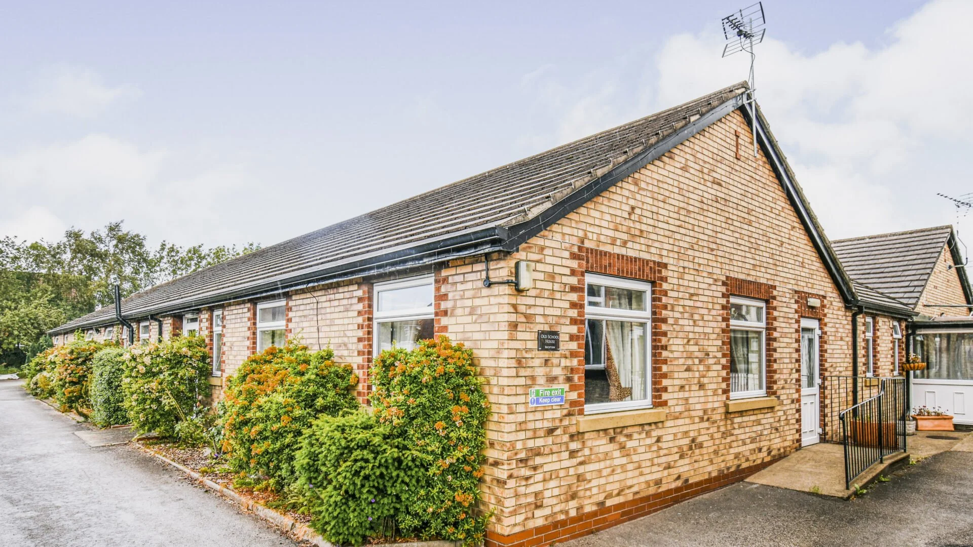 Exterior view of a brick residential building with several windows, surrounded by bushes and plants, under a cloudy sky.
