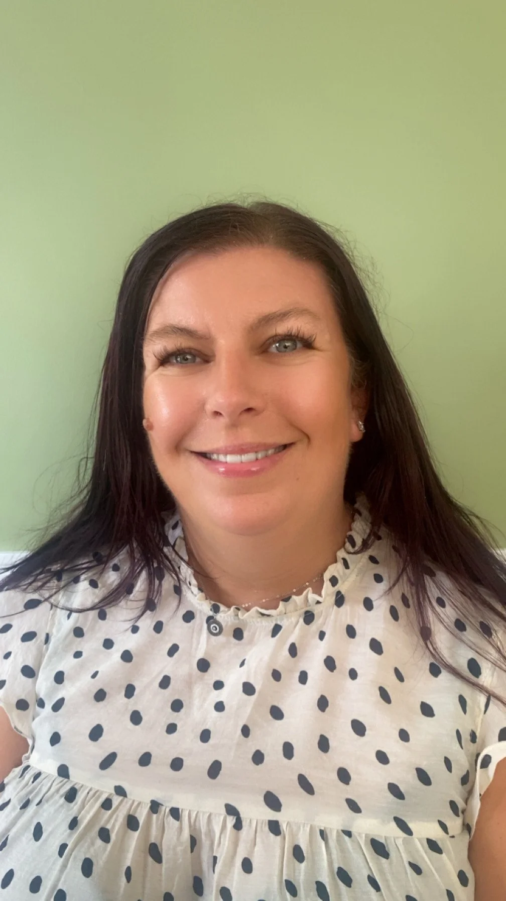 A smiling woman with dark hair and blue eyes posing in front of a light green wall, wearing a white dress with black polka dots.
