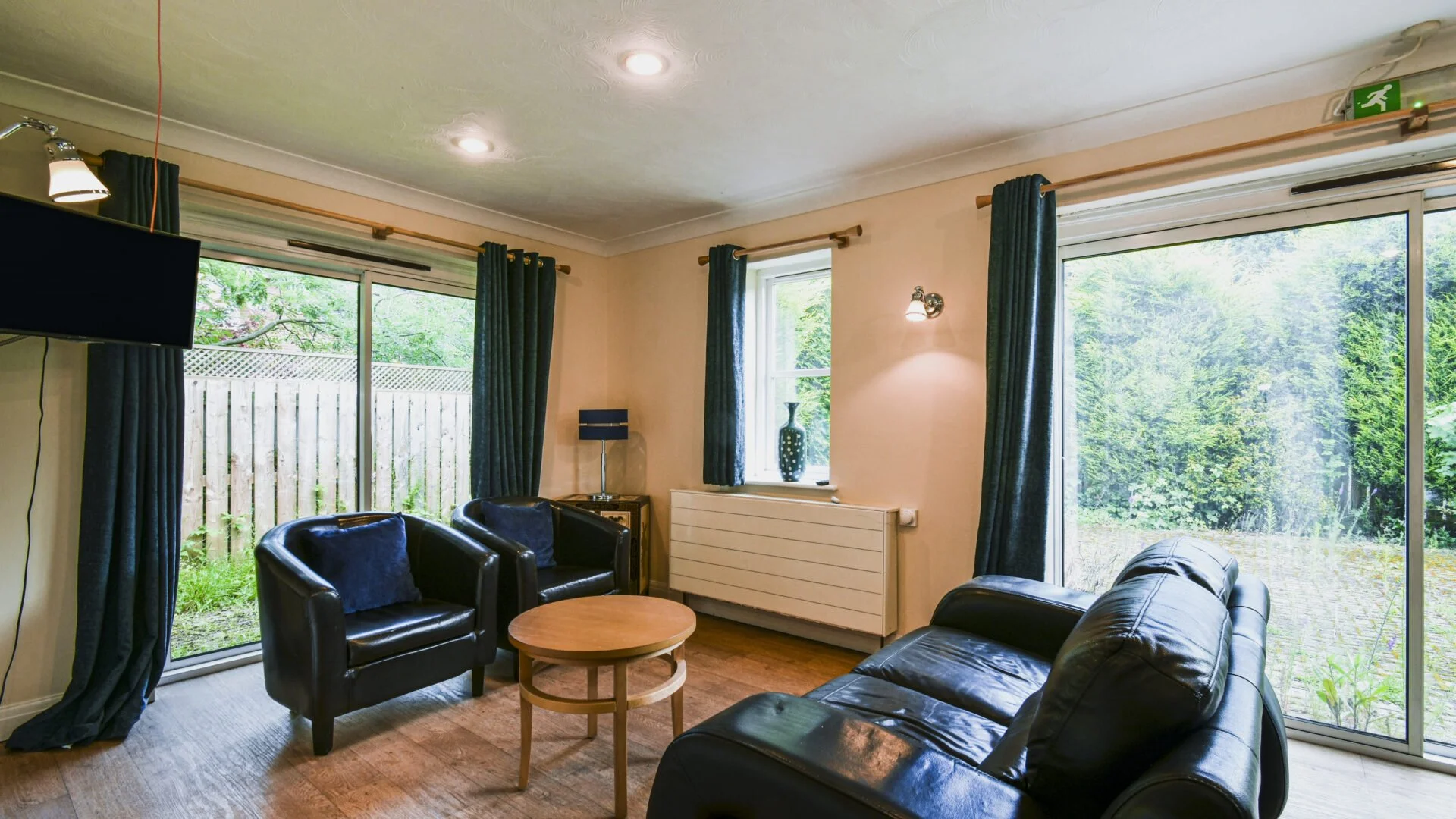 Living room with leather sofa, two armchairs, coffee table, flat-screen TV, Windows with blue curtains, and greenery outside.