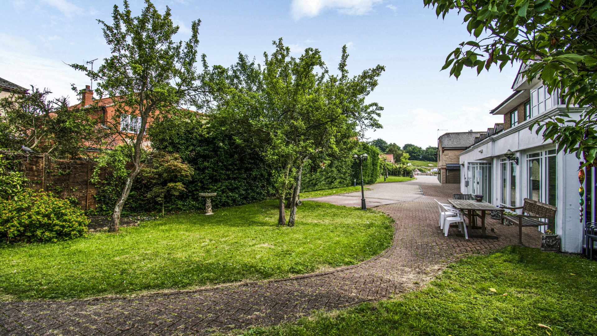A peaceful backyard with a brick path, green grass, trees, bushes, a birdbath, and outdoor seating near a white house with large windows.