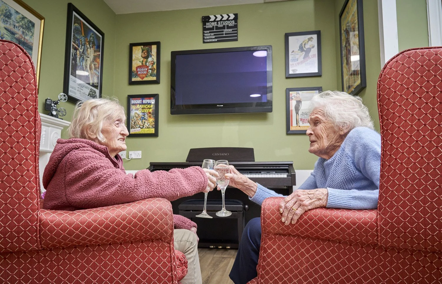 Two elderly women sitting in red armchairs, toasting with glasses of water in a cozy living room with framed movie posters on green walls.
