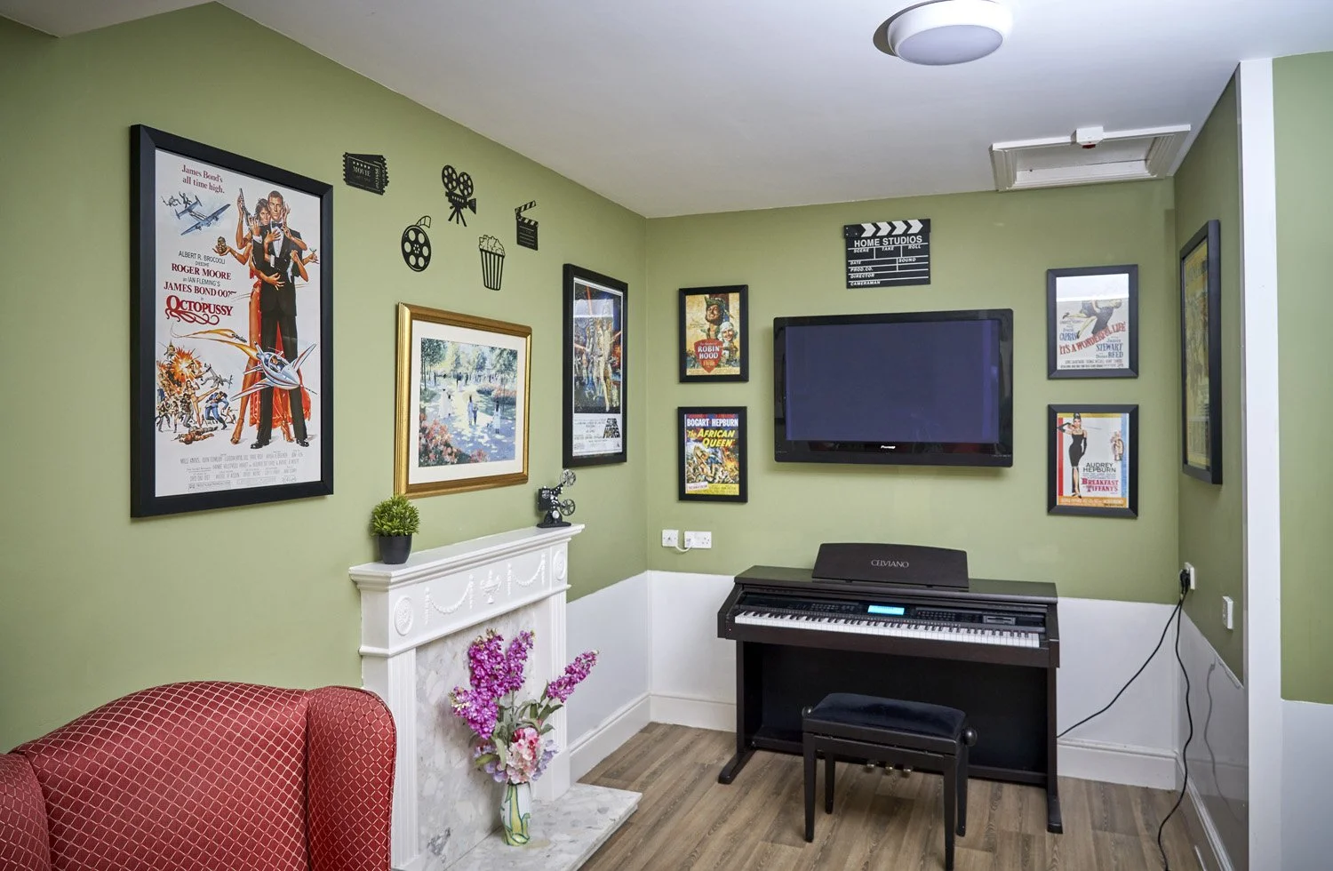Living room corner with green walls decorated with framed movie posters, a wall-mounted TV, a keyboard on a stand, and a red armchair. A small white fireplace with pink flowers in a green vase on top is also visible.