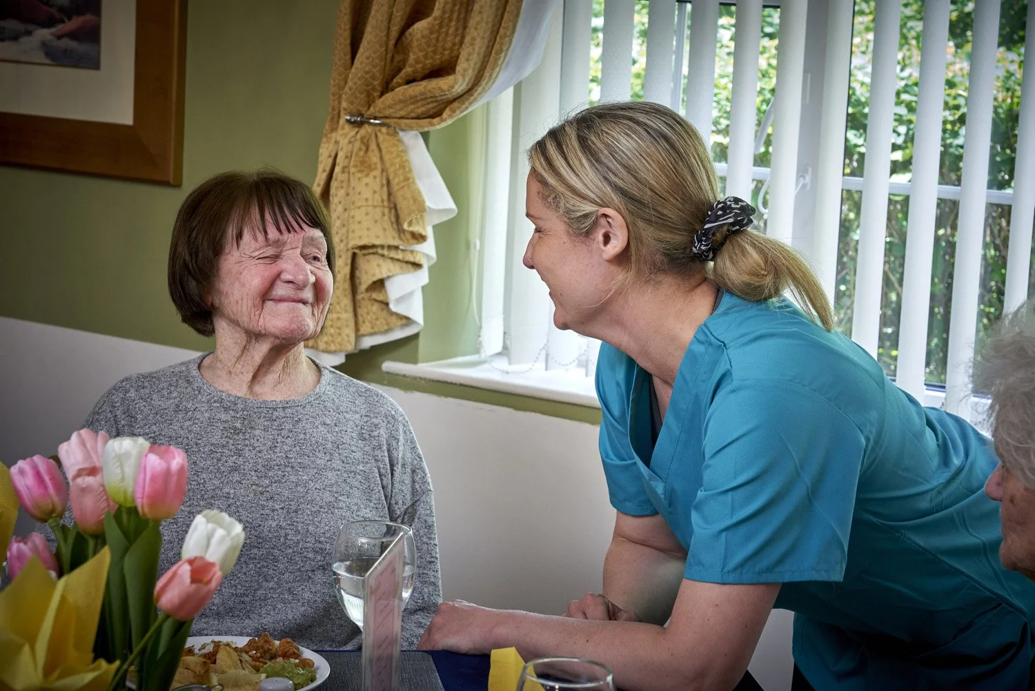 A woman in a blue uniform talking to an elderly woman at a dining table during a visit or celebration in a bright room with a window and floral decorations.