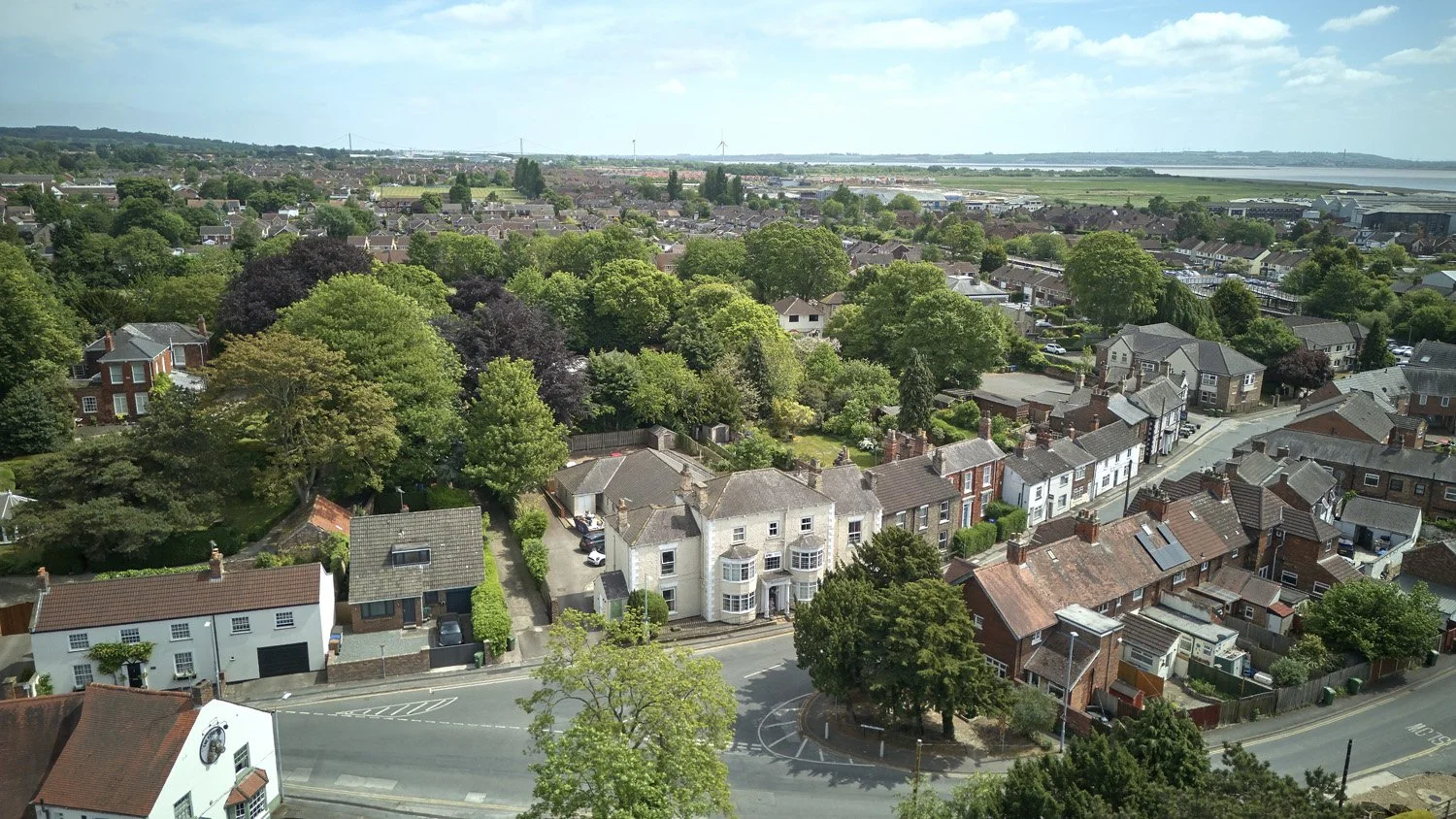 Aerial view of a residential neighborhood with a large green park and trees, various houses, and a body of water in the distance under a partly cloudy sky.