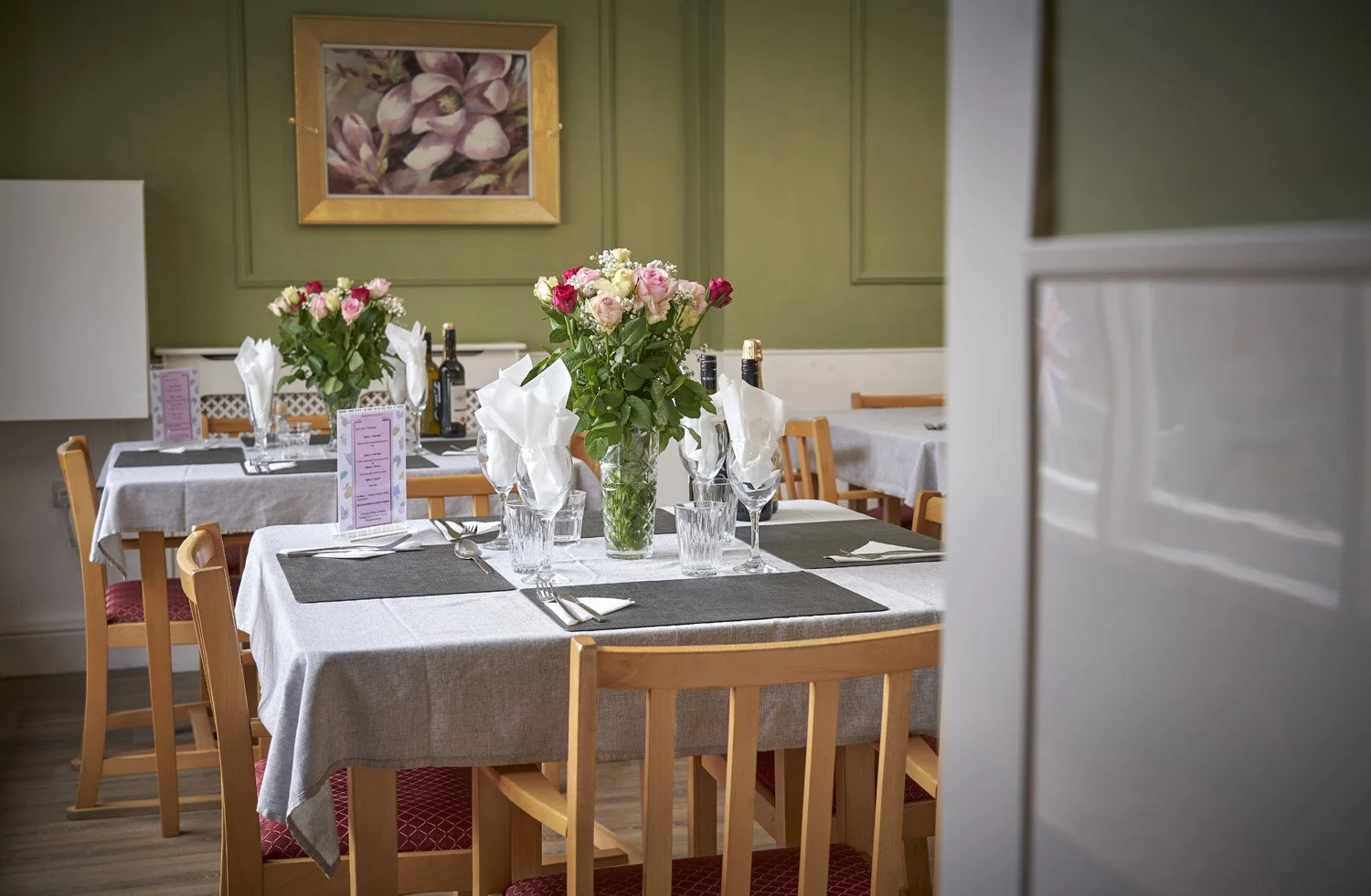 A set dining table decorated with pink and white roses in a vase, black placemats, silverware, wine bottles, folded napkins, and glasses, in a cozy restaurant with green walls and artwork.