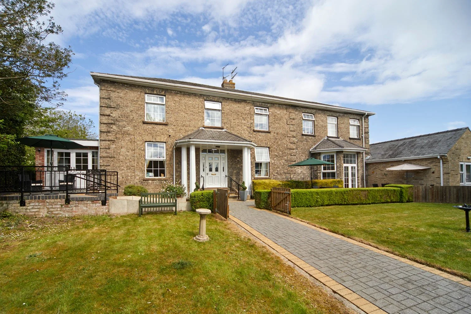 Front view of a brick house with a well-maintained lawn, paved walkway, and outdoor seating area, under a partly cloudy sky.