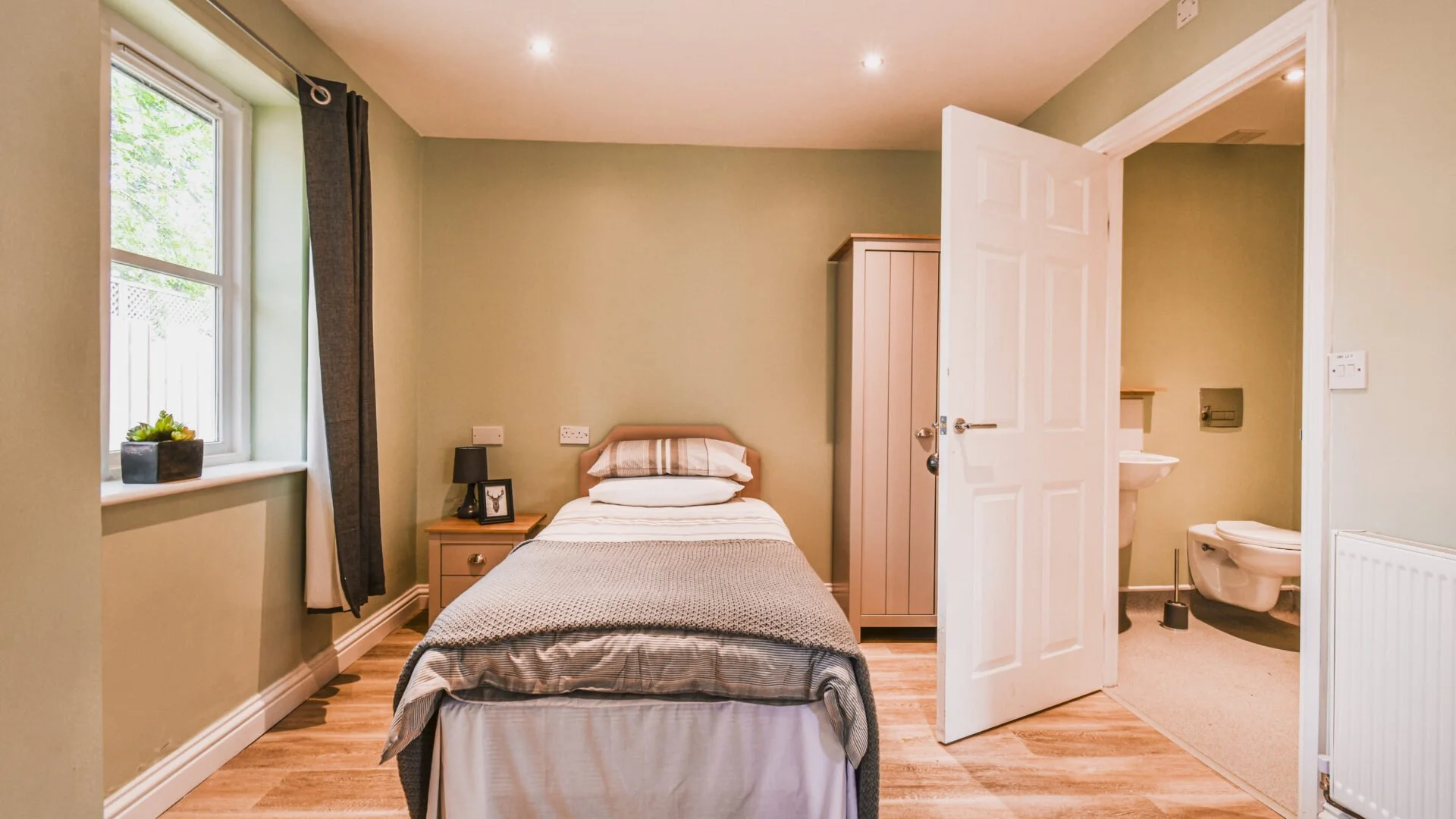 A small bedroom with a single bed, a nightstand, and a window with curtains. An open door reveals a bathroom with a toilet and sink.