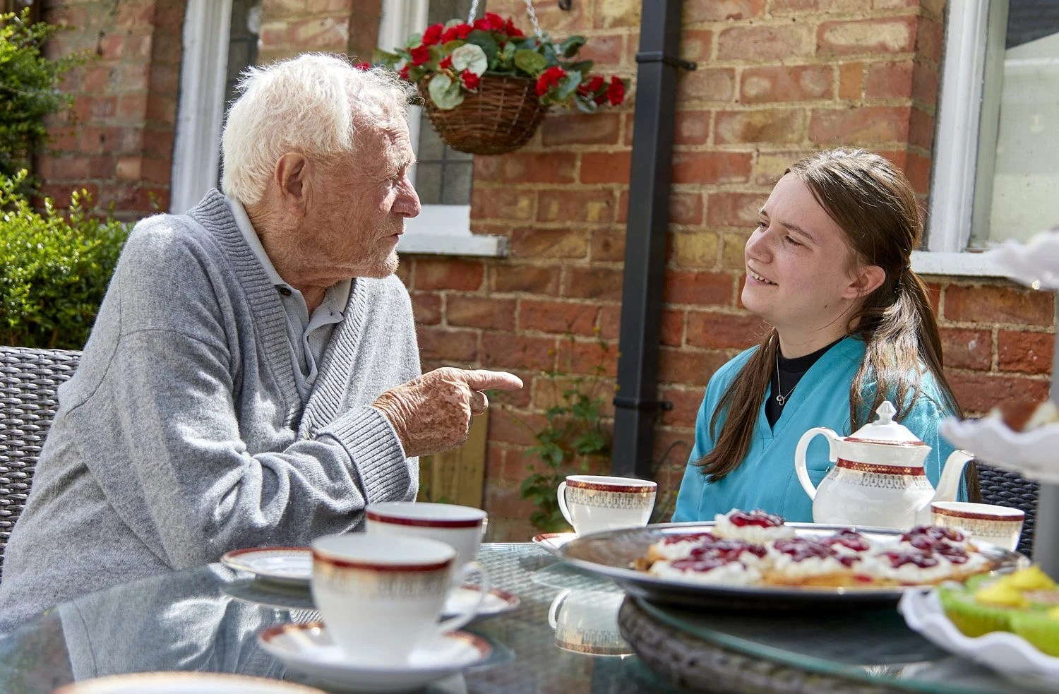 An elderly man with white hair and a gray sweater is pointing and speaking to a young woman with brown hair and a blue shirt, who is smiling. They are sitting at a table outdoors with a brick wall background, decorated with a hanging basket of red an
