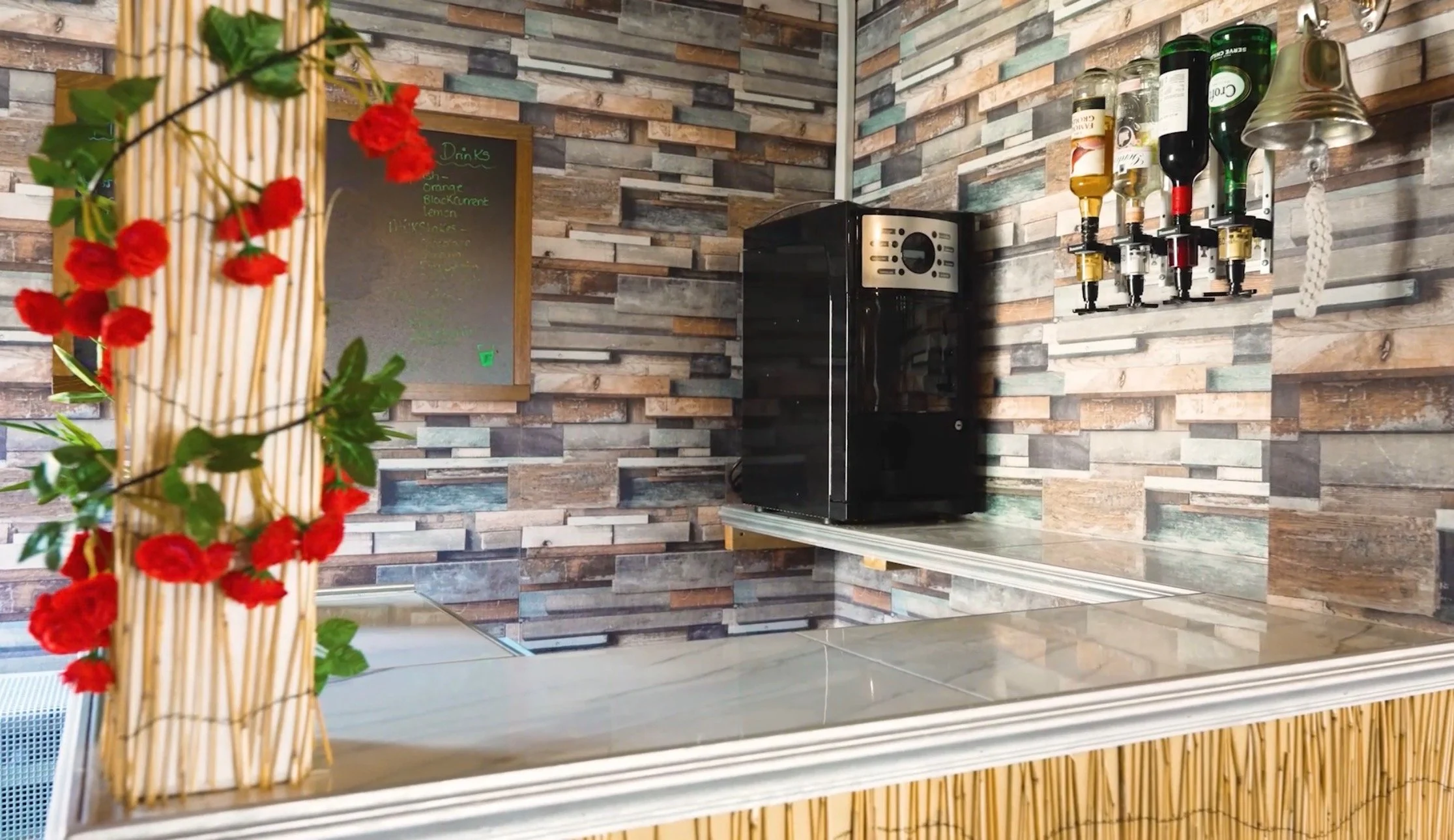 Decorated bar area with a flower arrangement on a counter, a black coffee machine, a wine rack with bottles, a small chalkboard menu, and a wooden wall background.