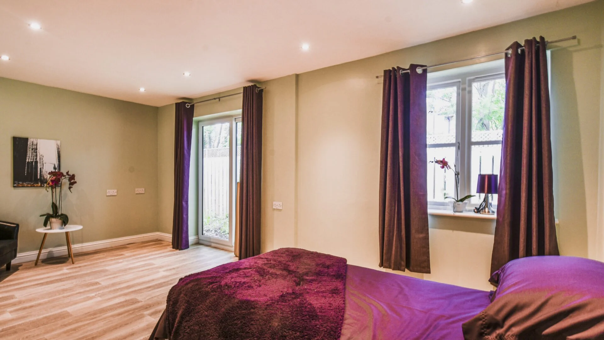 A bedroom with beige walls, wooden flooring, two windows with dark purple curtains. There is a bed with purple bedding and a blanket, a windowsill with a purple lamp and an orchid, a small white side table with an orchid, and a black armchair.