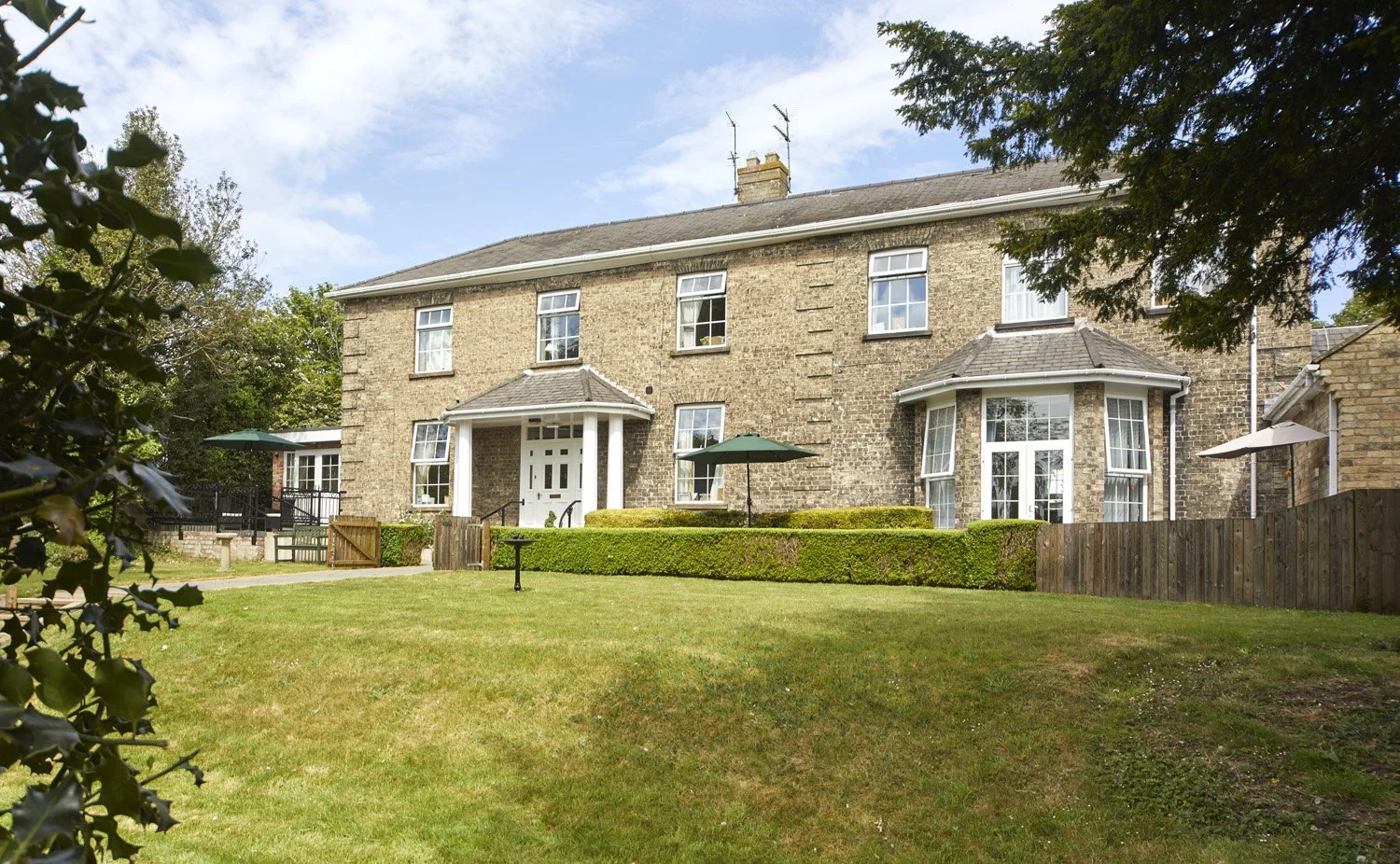 A large brick house with multiple windows and small porches, surrounded by a well-maintained lawn and garden with hedges, trees, and outdoor umbrellas, under a partly cloudy sky.