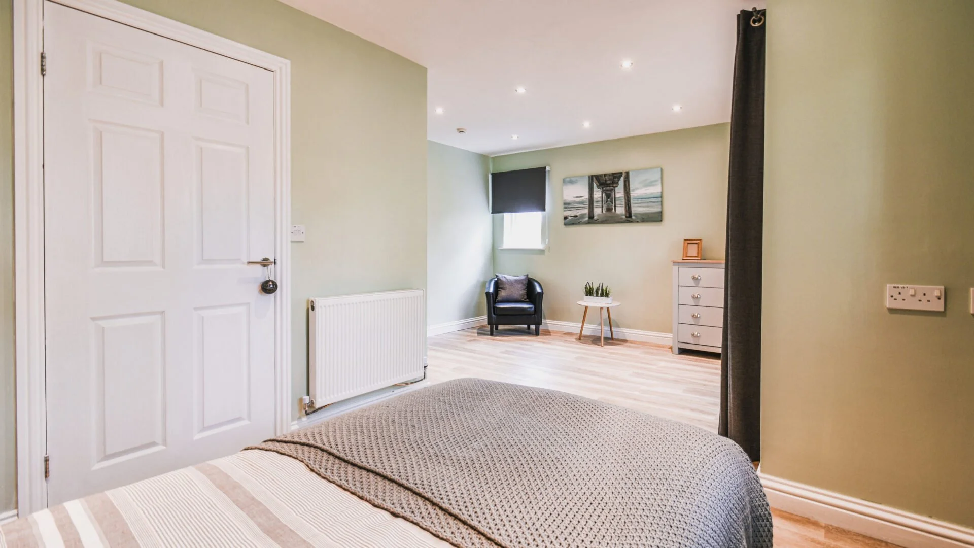 View from a bedroom into a sitting area with a black armchair, small table with a plant, dresser, and a window with a rolled-up blind, green walls, wooden flooring, and ceiling lights.
