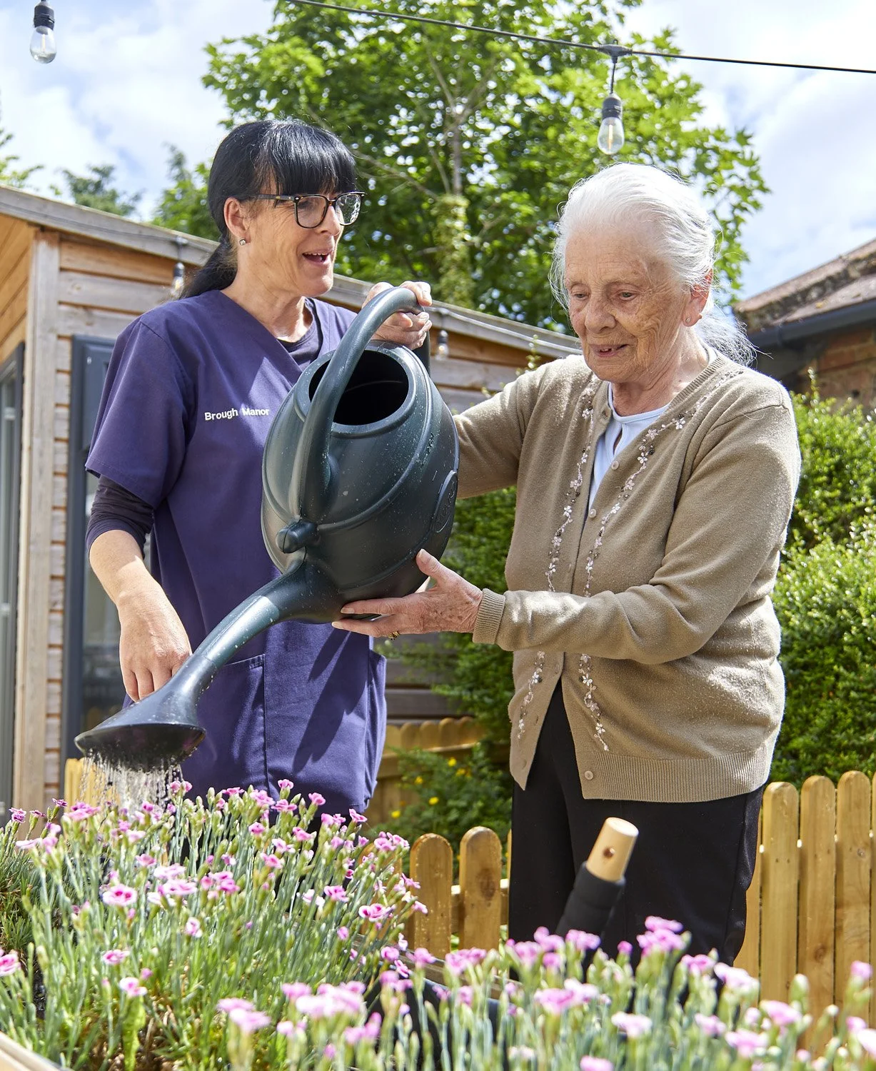 A caretaker and an elderly woman watering plants in a garden.