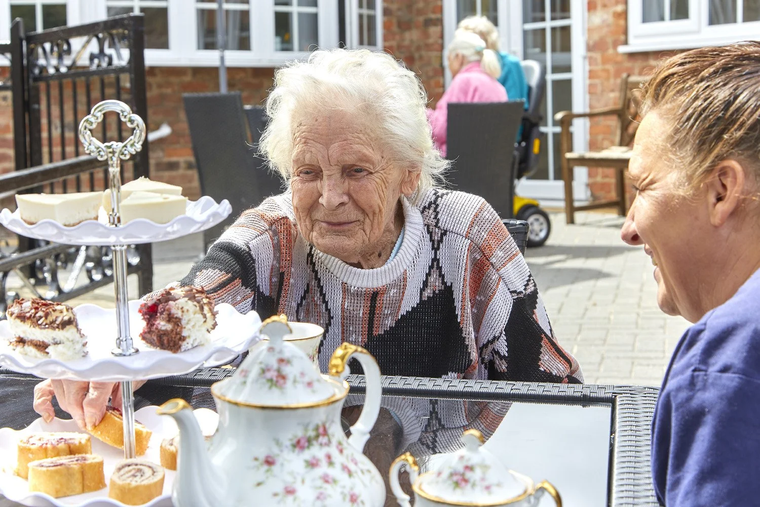 Older woman and younger woman sharing tea and desserts during outdoor tea party, with a tiered tray of cakes and teapot on table.