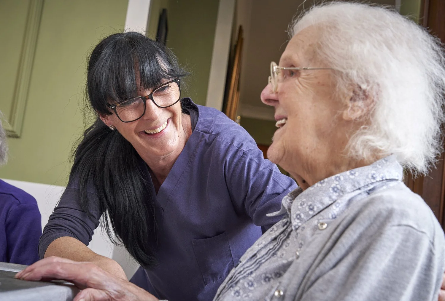 Nurse smiling and talking to an elderly woman with white hair, sitting together indoors.