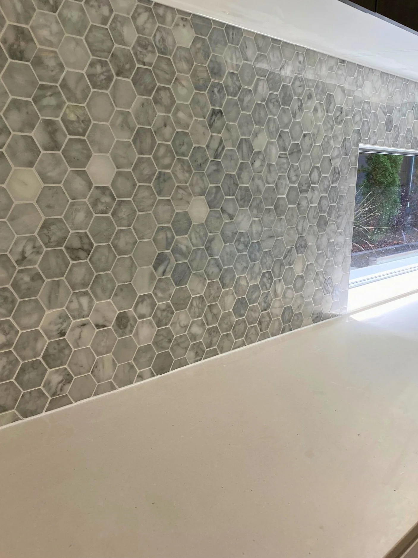 A kitchen backsplash with hexagonal marble tiles in gray and white tones above a beige countertop, with a window showing outdoor vegetation.