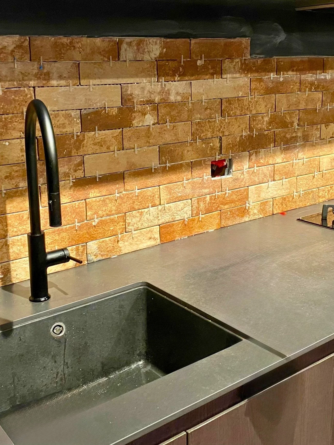Kitchen sink with black faucet, unfinished brick wall with tiles being installed, and a countertop with an electrical outlet box.