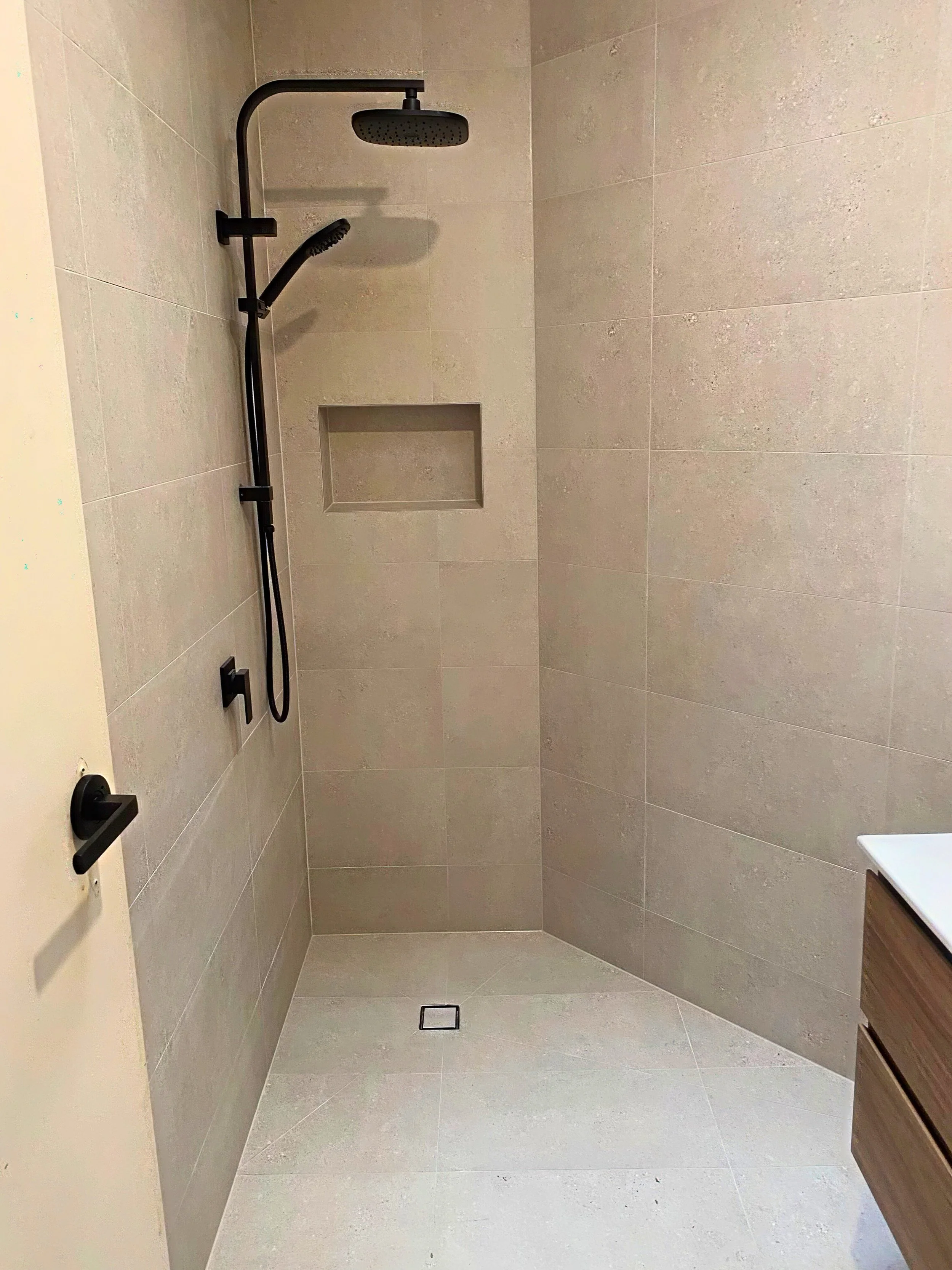 Modern walk-in shower with beige tile walls, rainfall showerhead, detachable shower wand, built-in niche, and a small square drain on the floor.
