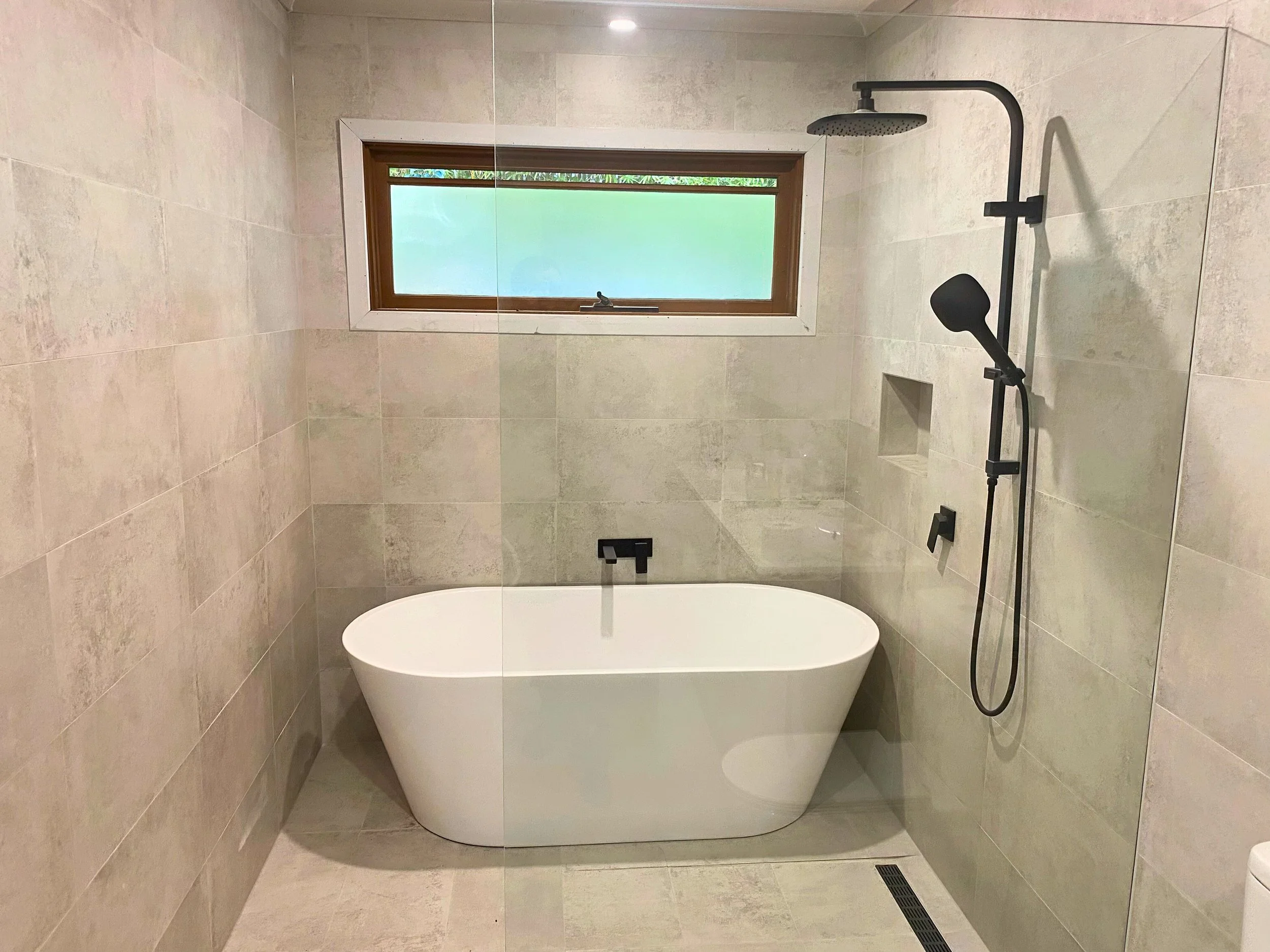 Modern bathroom with a white freestanding bathtub, black wall-mounted shower fixtures, a rectangular window with frosted glass, beige tiled walls and floor, and a small built-in shower niche.