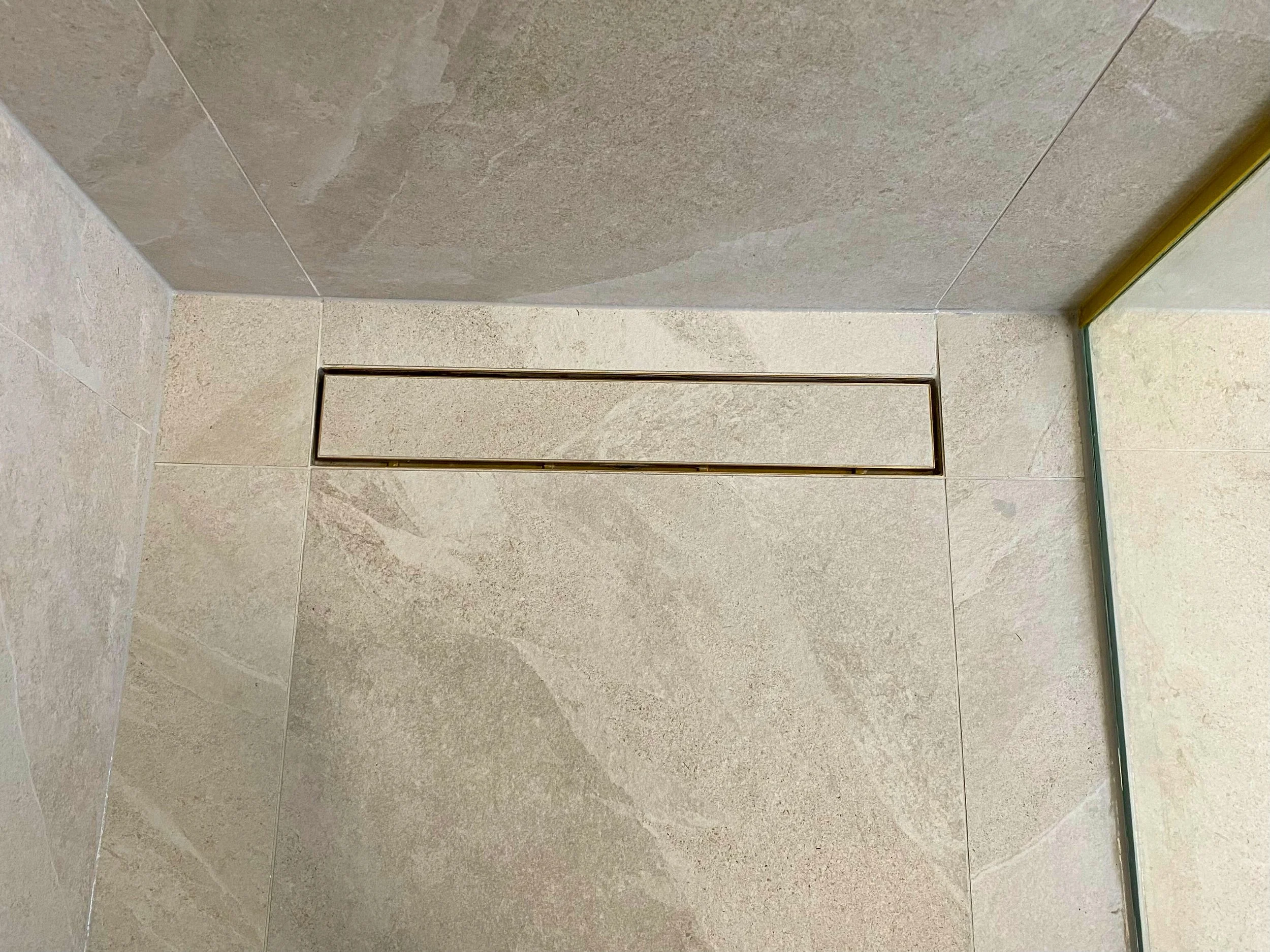 Close-up of a beige tiled shower wall with a rectangular metal drain cover and a glass panel on the right side.