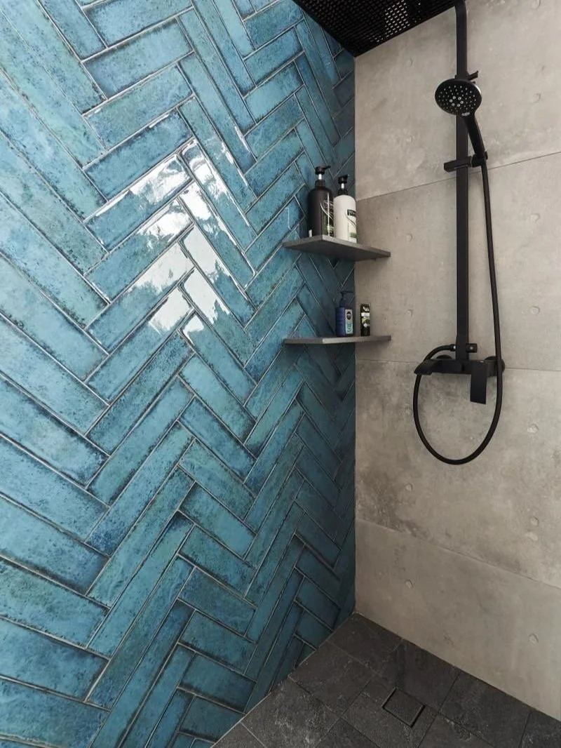 Shower area with a black rain shower head, a handheld shower, and two gray shelves holding toiletries, against a wall with blue herringbone tiles and concrete tiles.